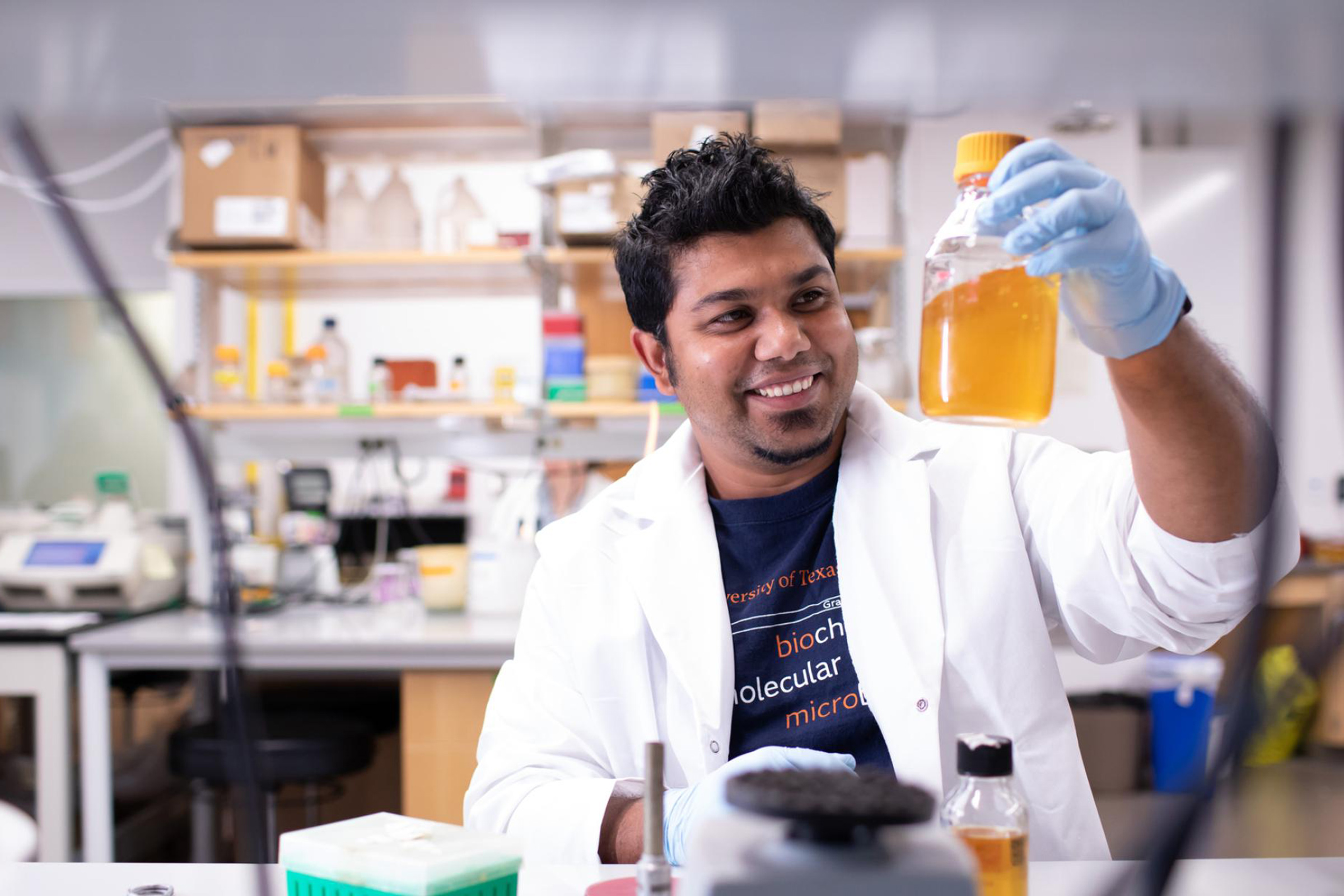 A graduate student in a white lab coat and PPE smiles at a container he holds up to view