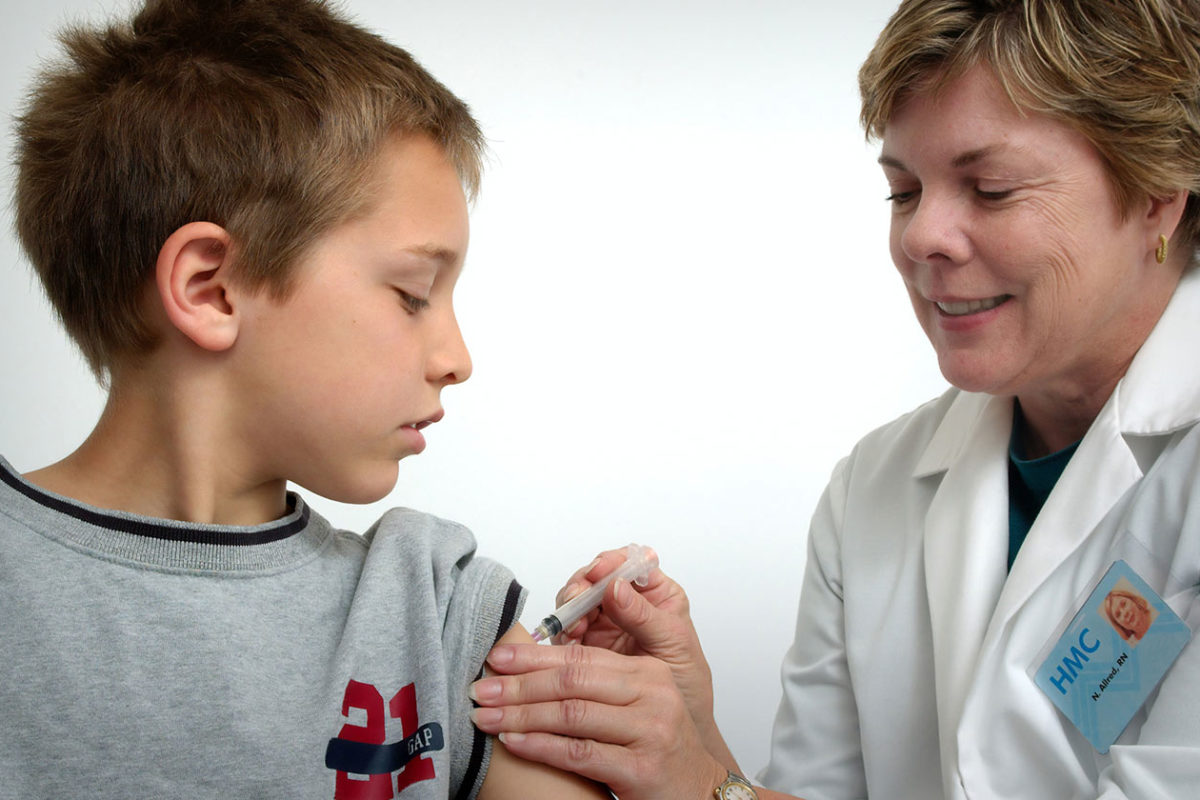 A young boy on the left receives a vaccination in his arm from a woman in a lab coat on the right 