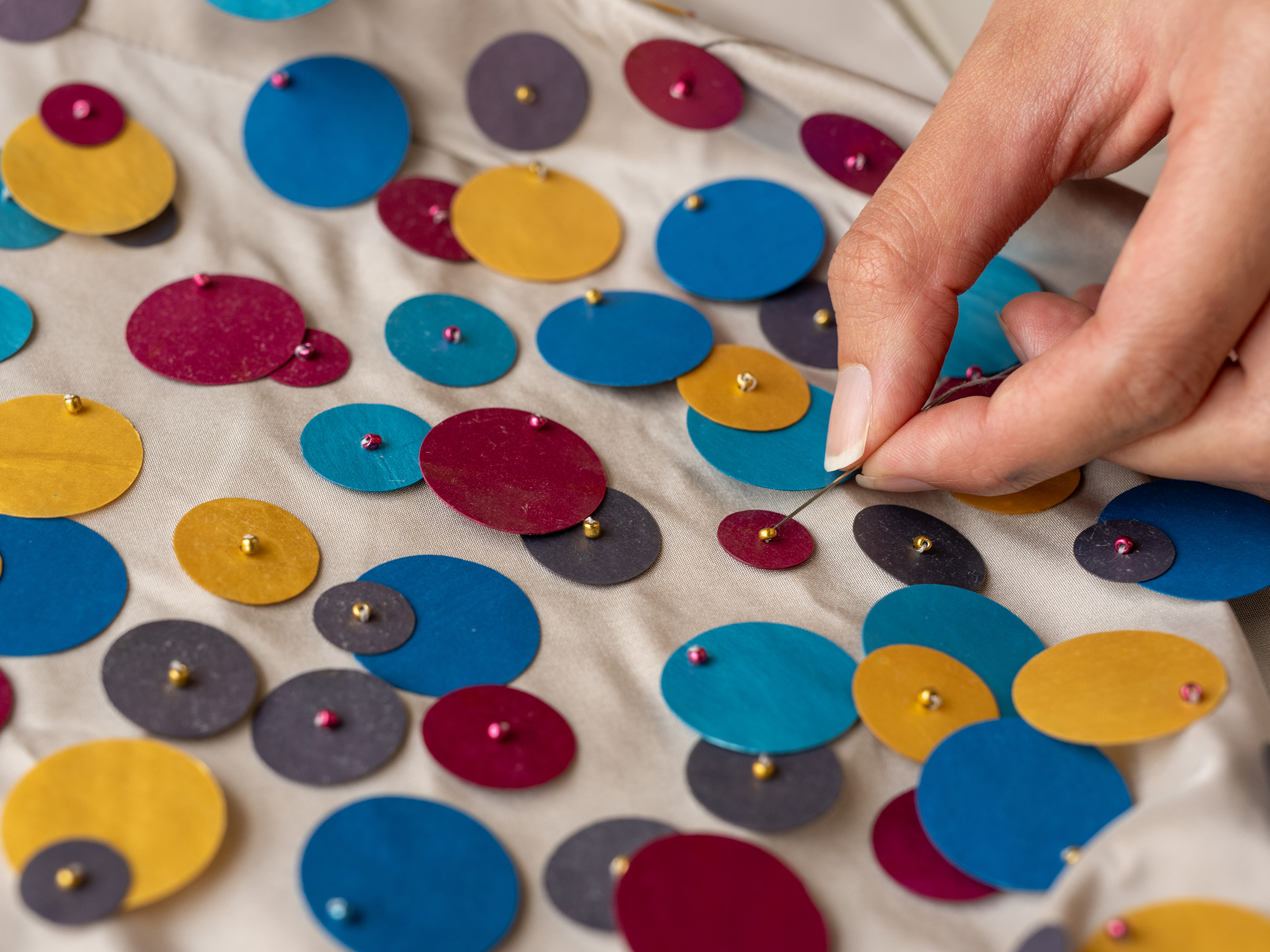 A hand sews sustainable sequins onto a piece of fabric.