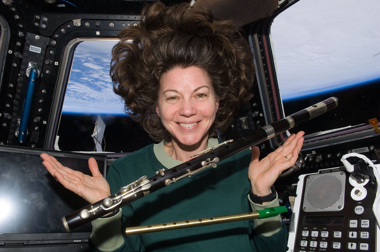 A space station astronaut holds her hands up in a capsule in space while musical instruments and her long hair float about in zero gravity. The Earth can be seen out the sindwo behidn ehr.
