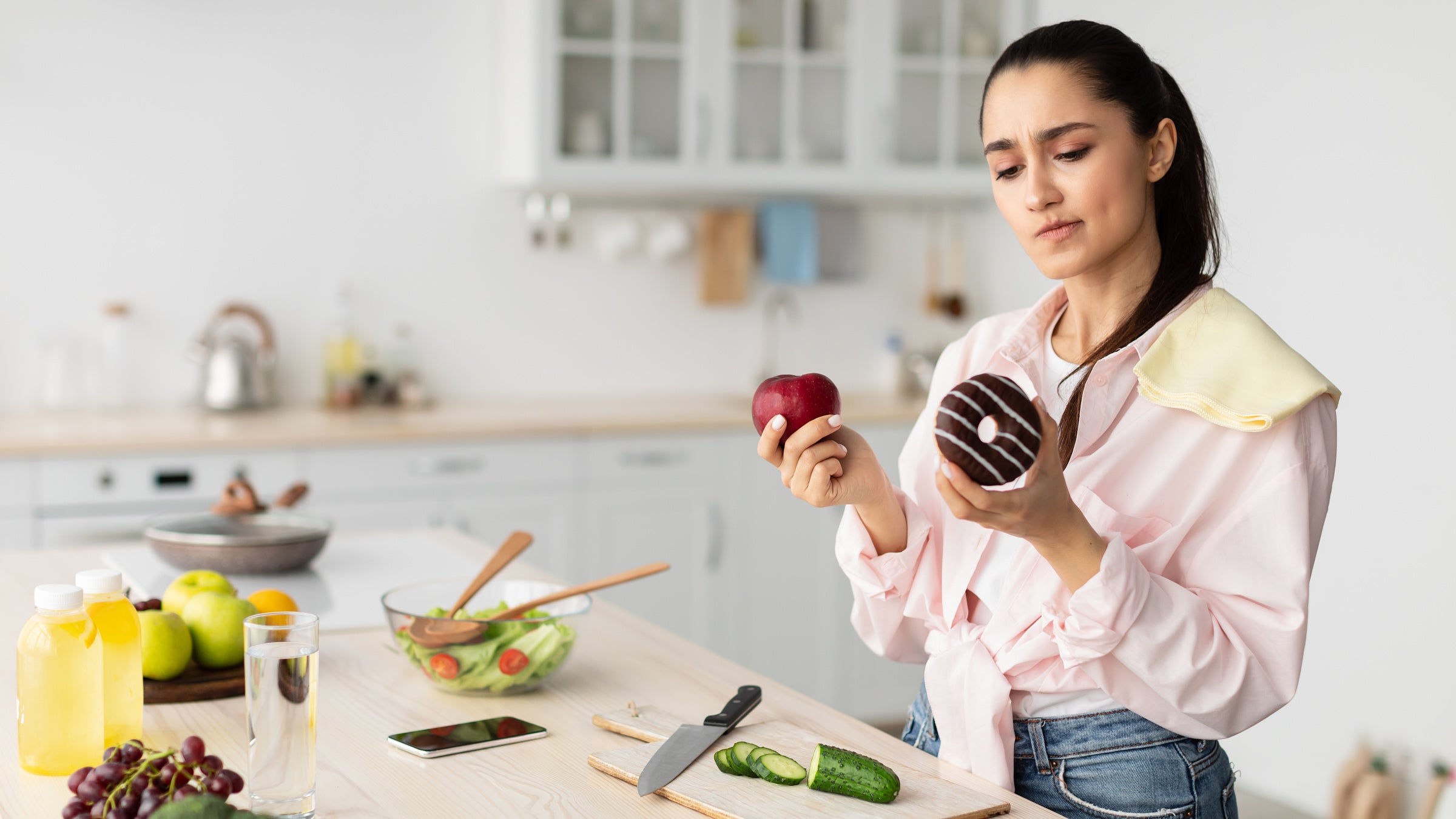 A woman stands in a kitchen holding an apple in one hand and a donut in the other, debating which one to eat