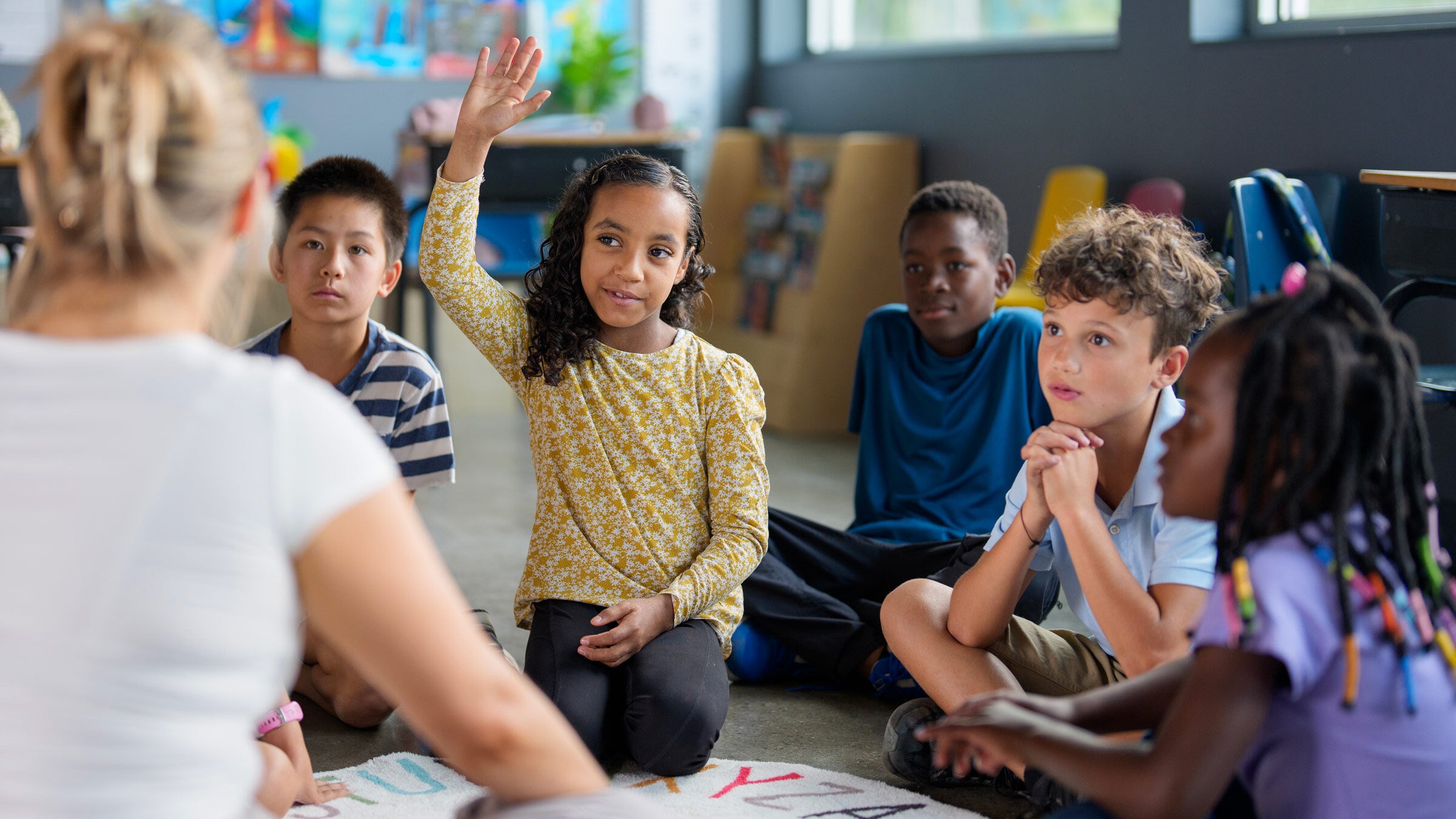 Several young children sit in front of a teacher whose back is to the viewer. One girl is raising her hand to ask a question.