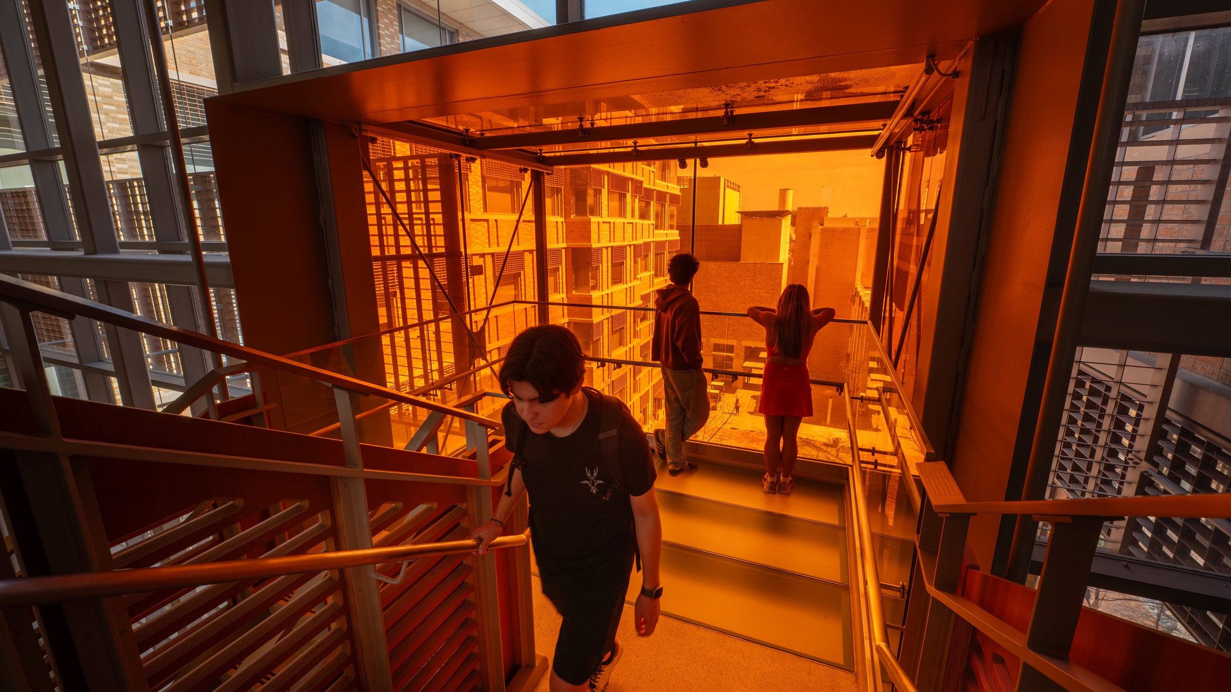 Two students view the UT campus out an upper-floor, burnt orange oriel while another student ascends stairs