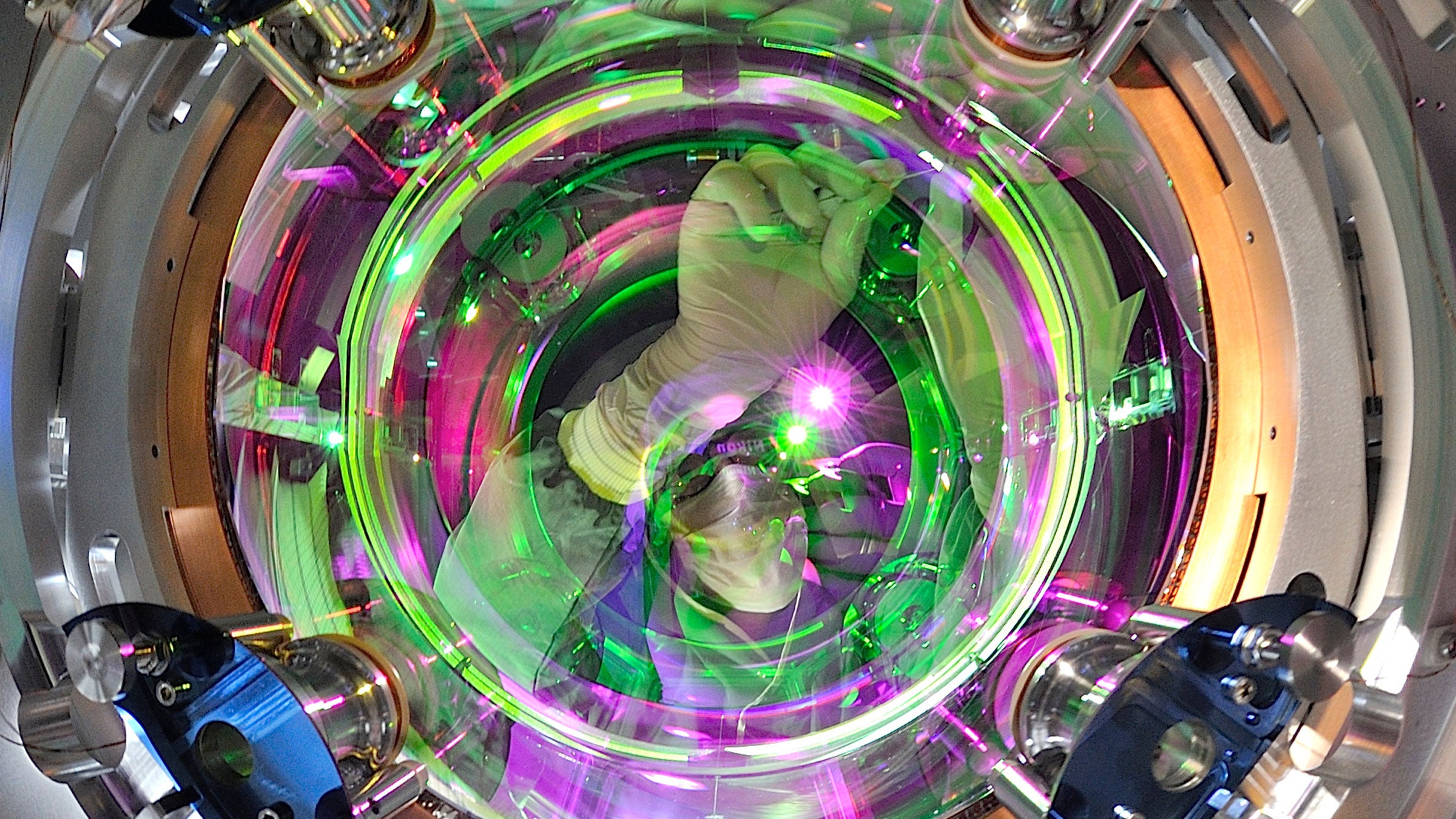 A technician wearing a facemask makes adjustments to a piece of scientific equipment. The person is visible through a round glass window surrounded by metal and regions lit by purple and green light.