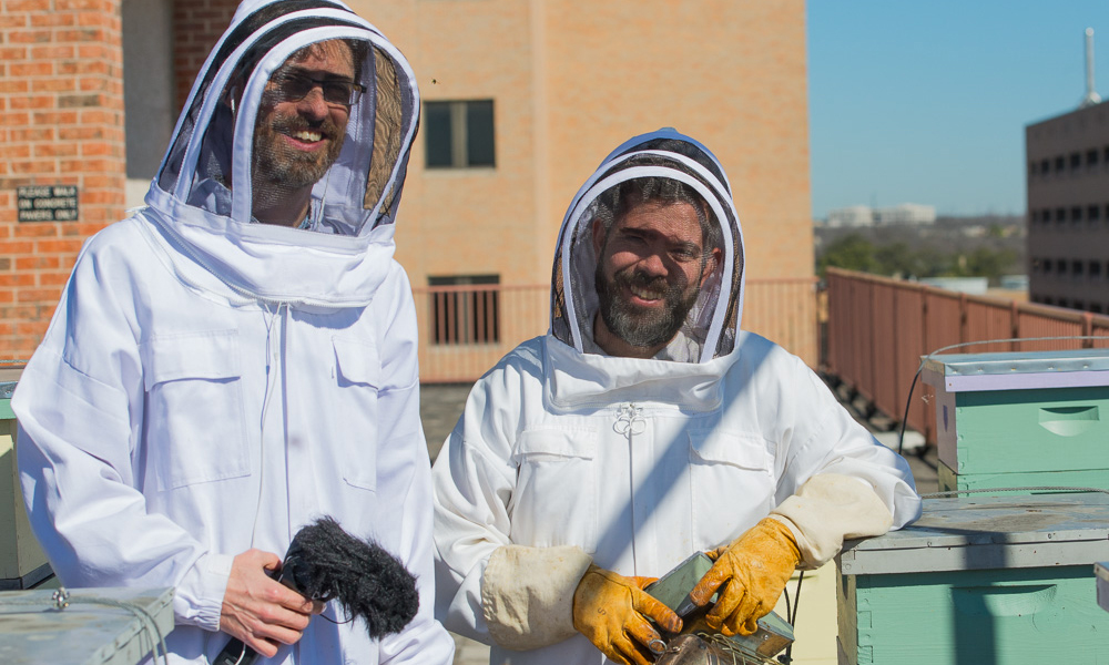 Two men in white bee suits with mesh helmets pose on the roof of a building