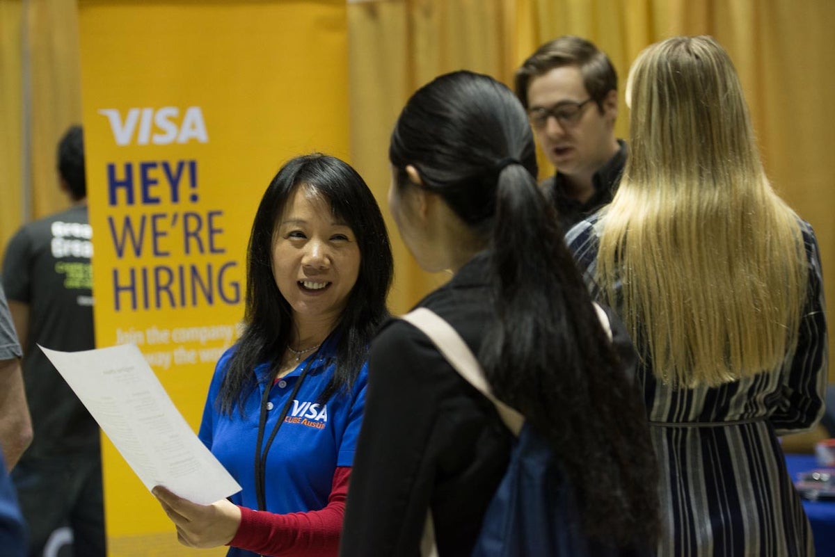 A woman in a Visa shirt stands in front of a banner that reads: "Visa, Hey! We're Hiring" while talking with a job candidate at a career fair and reviewing her resume