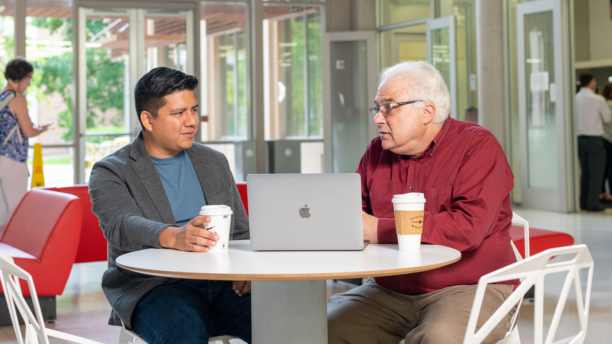 Two men at a table in a public cafe having a conversation with coffee cups and a laptop computer