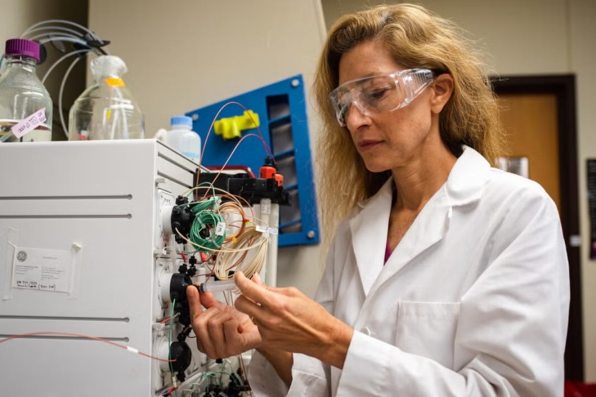 A woman in a lab coat and goggles connects a plastic tube to a piece of laboratory equipment laden with wires.