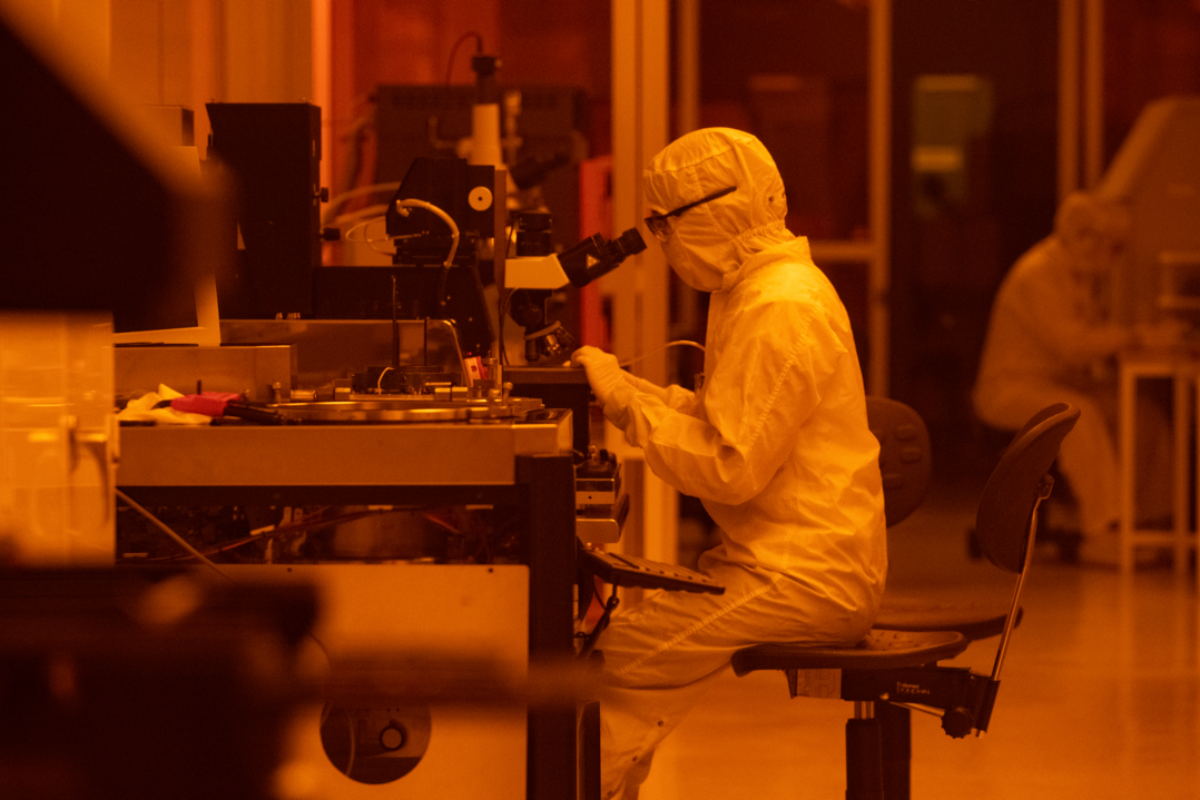 A person in a white full body clean room suit peers into a microscope