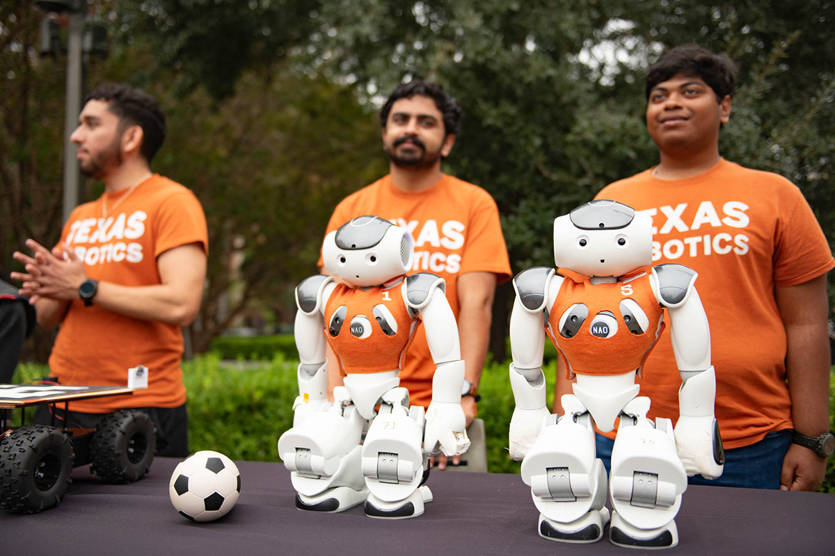Two small white robots crouch next to a tiny soccer ball on a table in front of students wearing orange shirts that say Texas Robotics