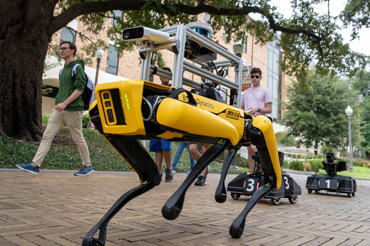 A yellow robot with four legs parades down Speedway with rolling robots in its wake as students look on