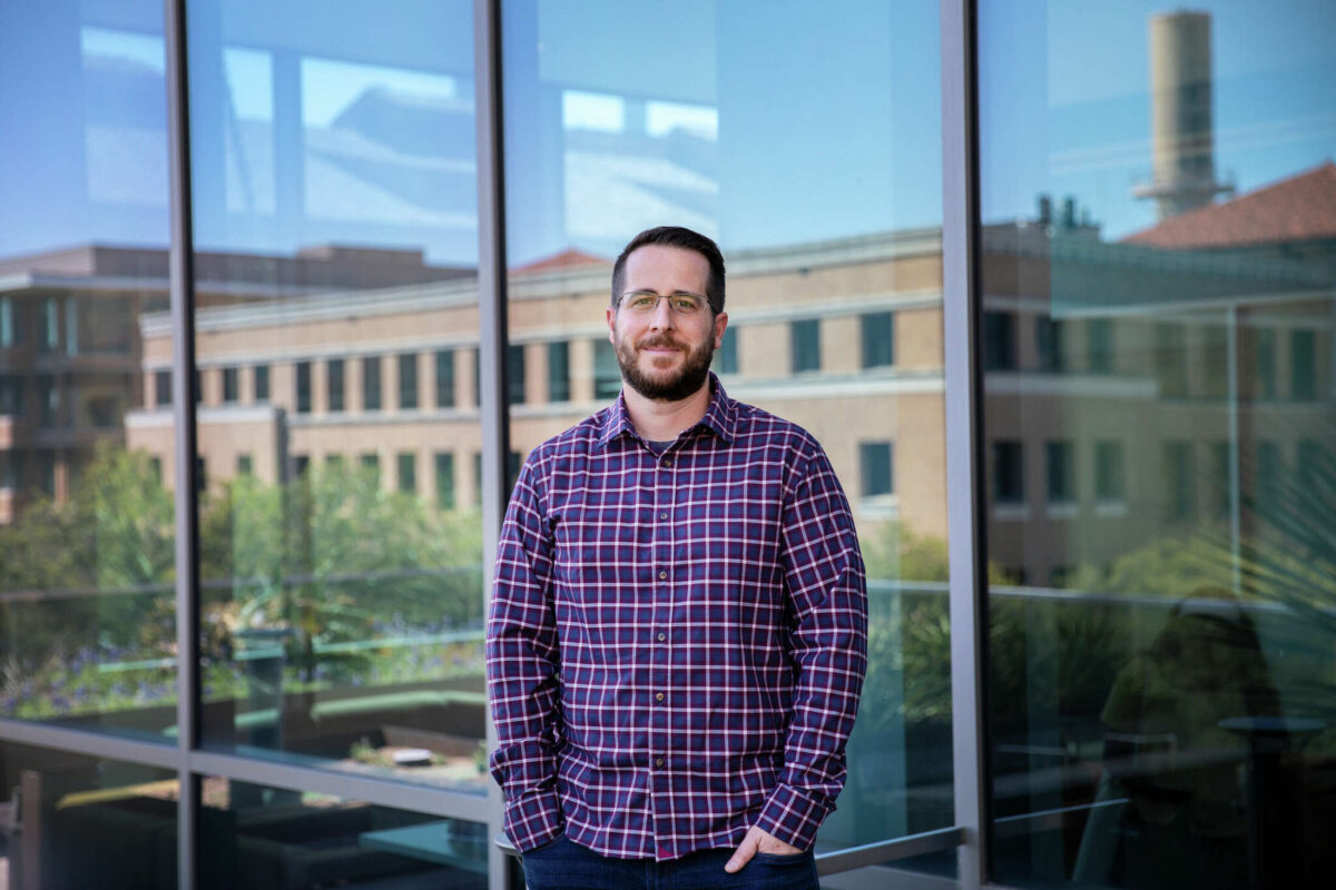 Jason McLellan stands with hands in pockets in front of Norman Hackerman Building's glass walls as other campus buildings and foliage appear in the reflection behind him