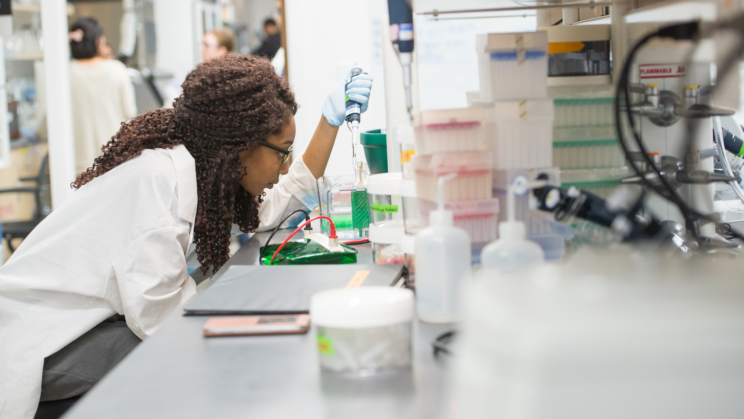 A student researcher pipettes in a lab wearing a lab coat