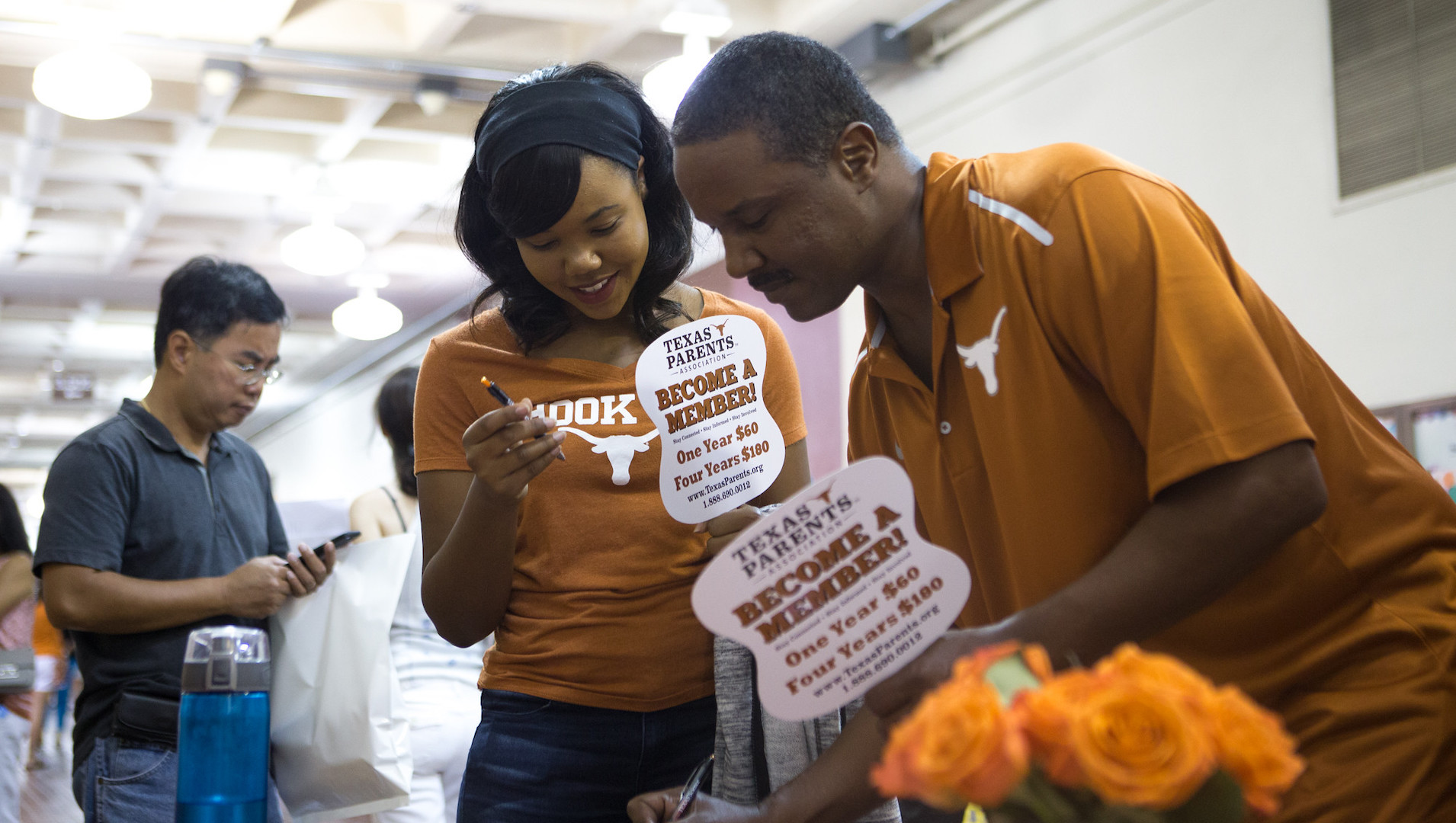 Family members in burnt orange shirts with Longhorns examine things at a table with signs that encourage people to become a member of Texas Parents
