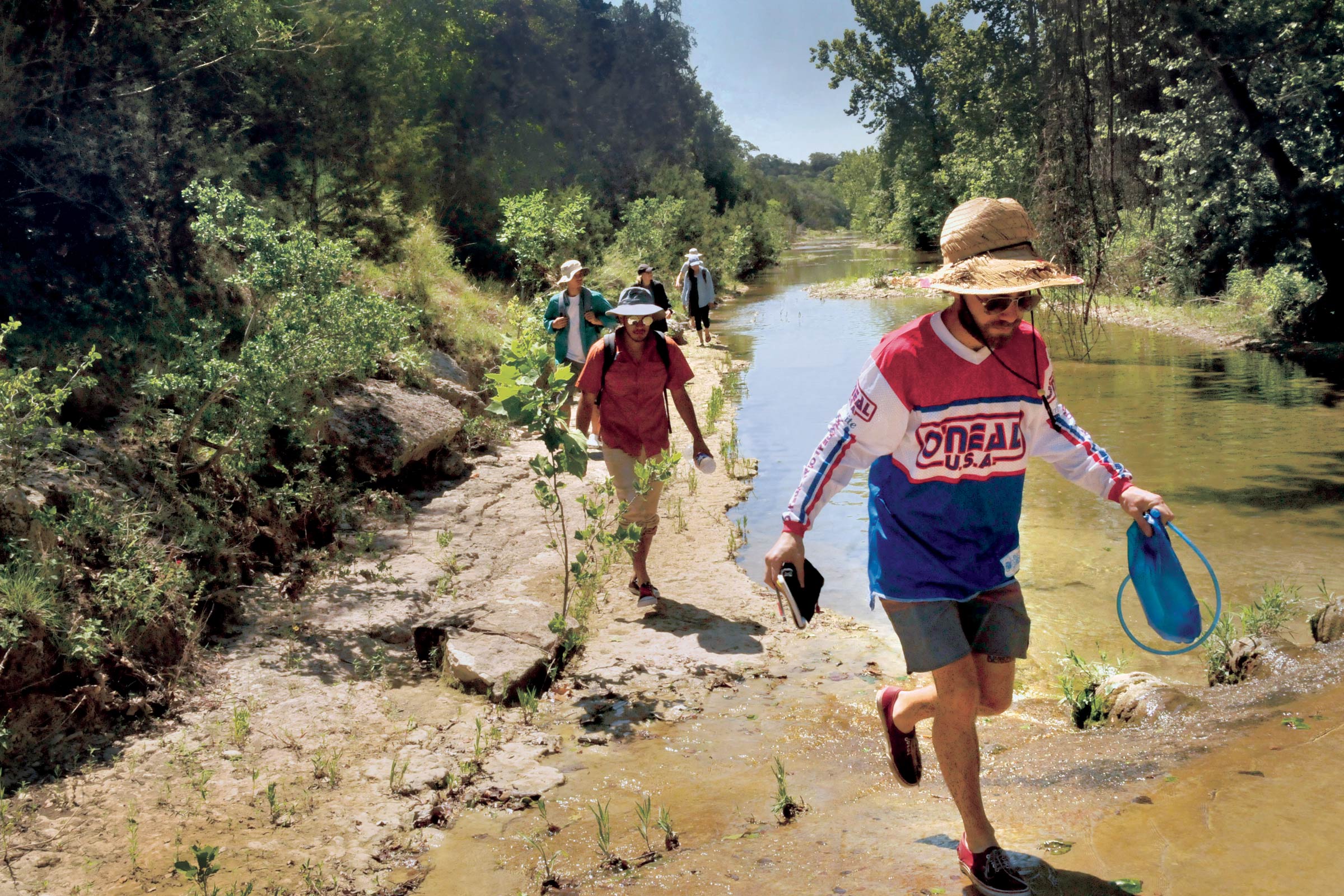 A young man in a hat holding equipment leads a group of students down a creek at a Hill Country-based field station