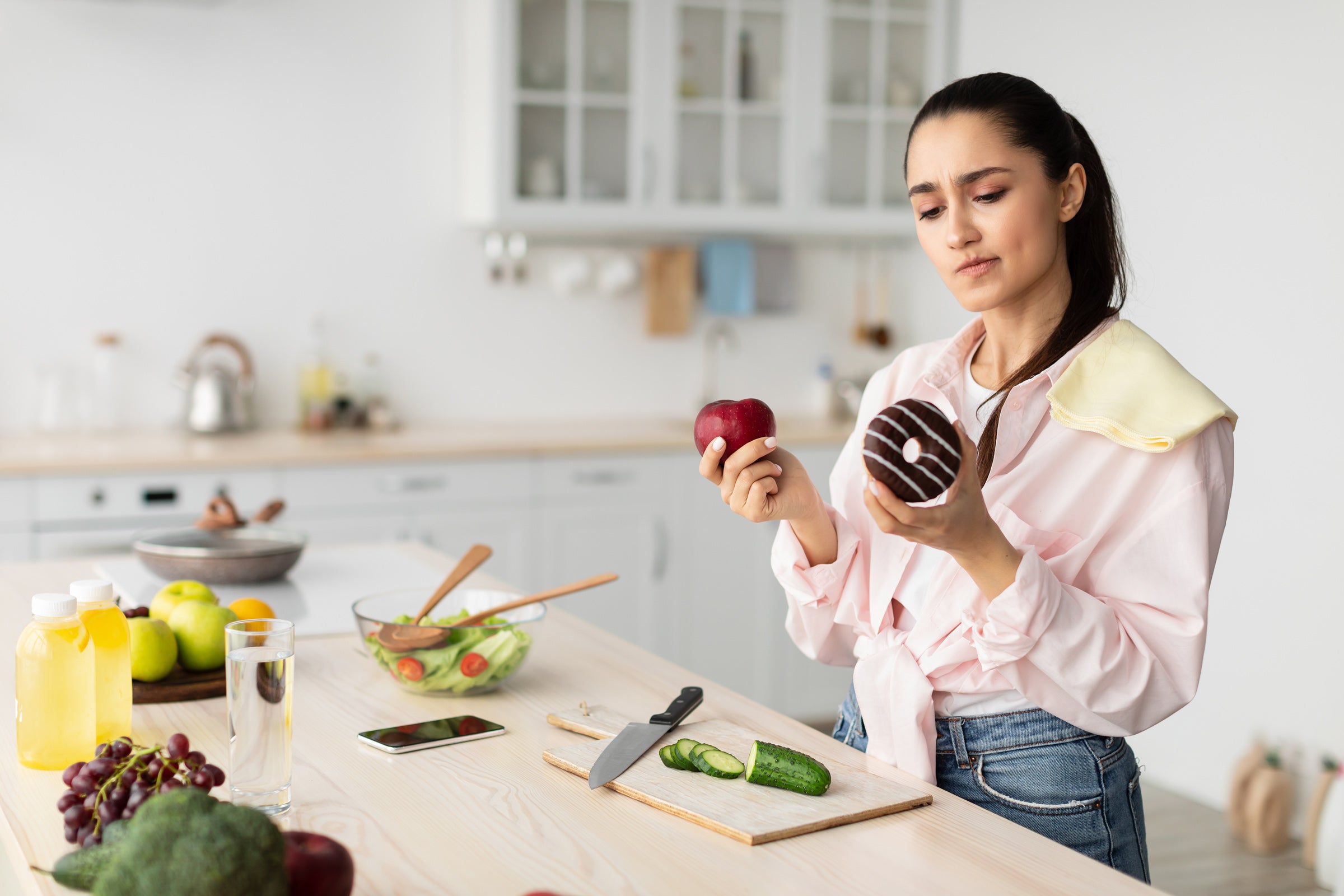 A woman stands in a kitchen holding an apple in one hand and a donut in the other, debating which one to eat