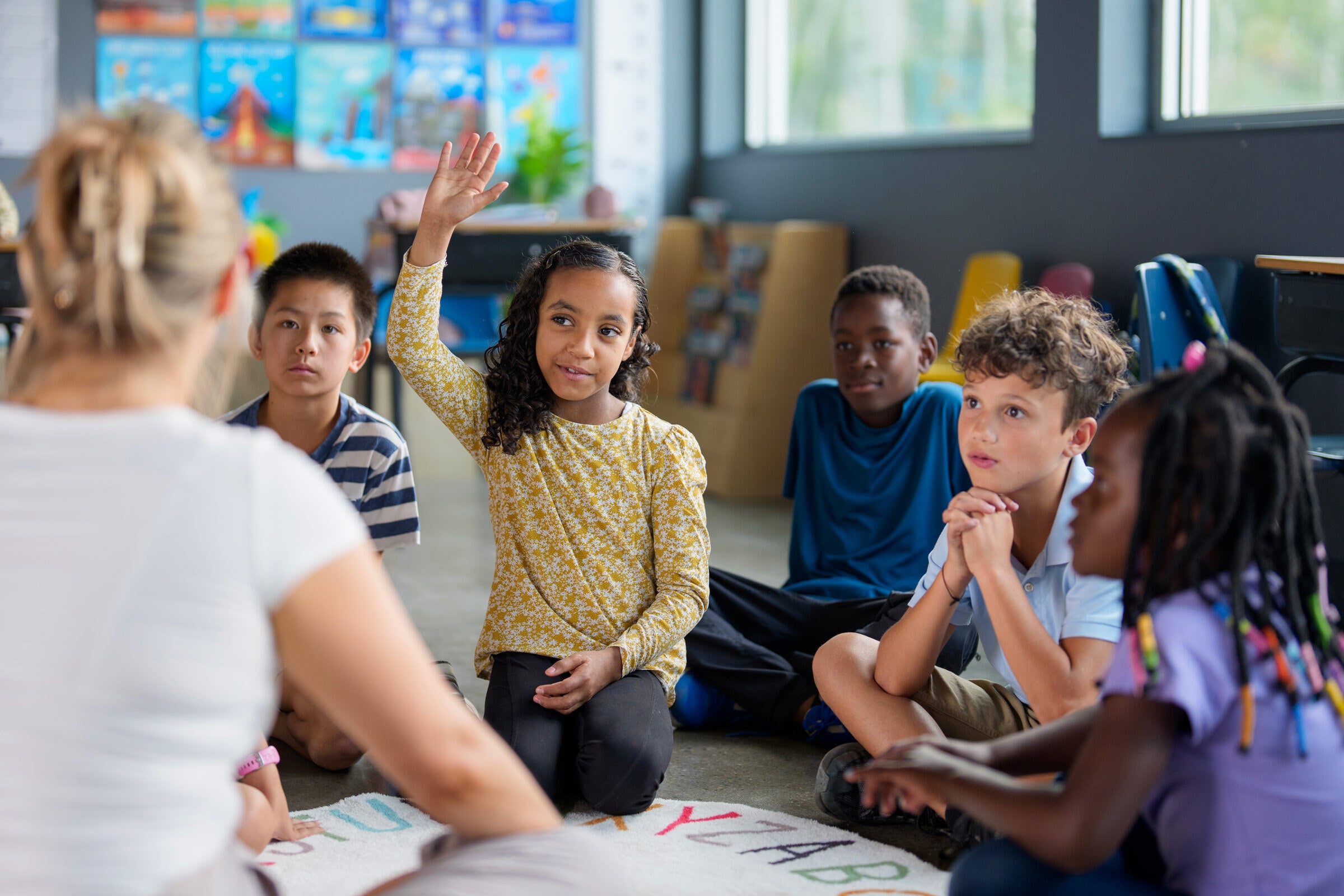 Several young children sit in front of a teacher whose back is to the viewer. One girl is raising her hand to ask a question.