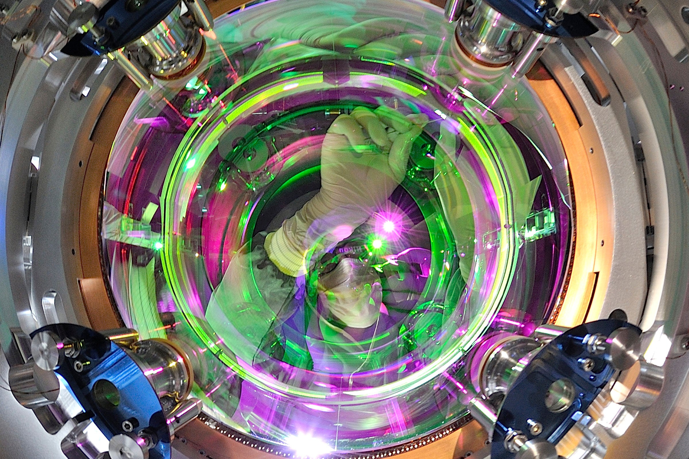 A technician wearing a facemask makes adjustments to a piece of scientific equipment. The person is visible through a round glass window surrounded by metal and regions lit by purple and green light.