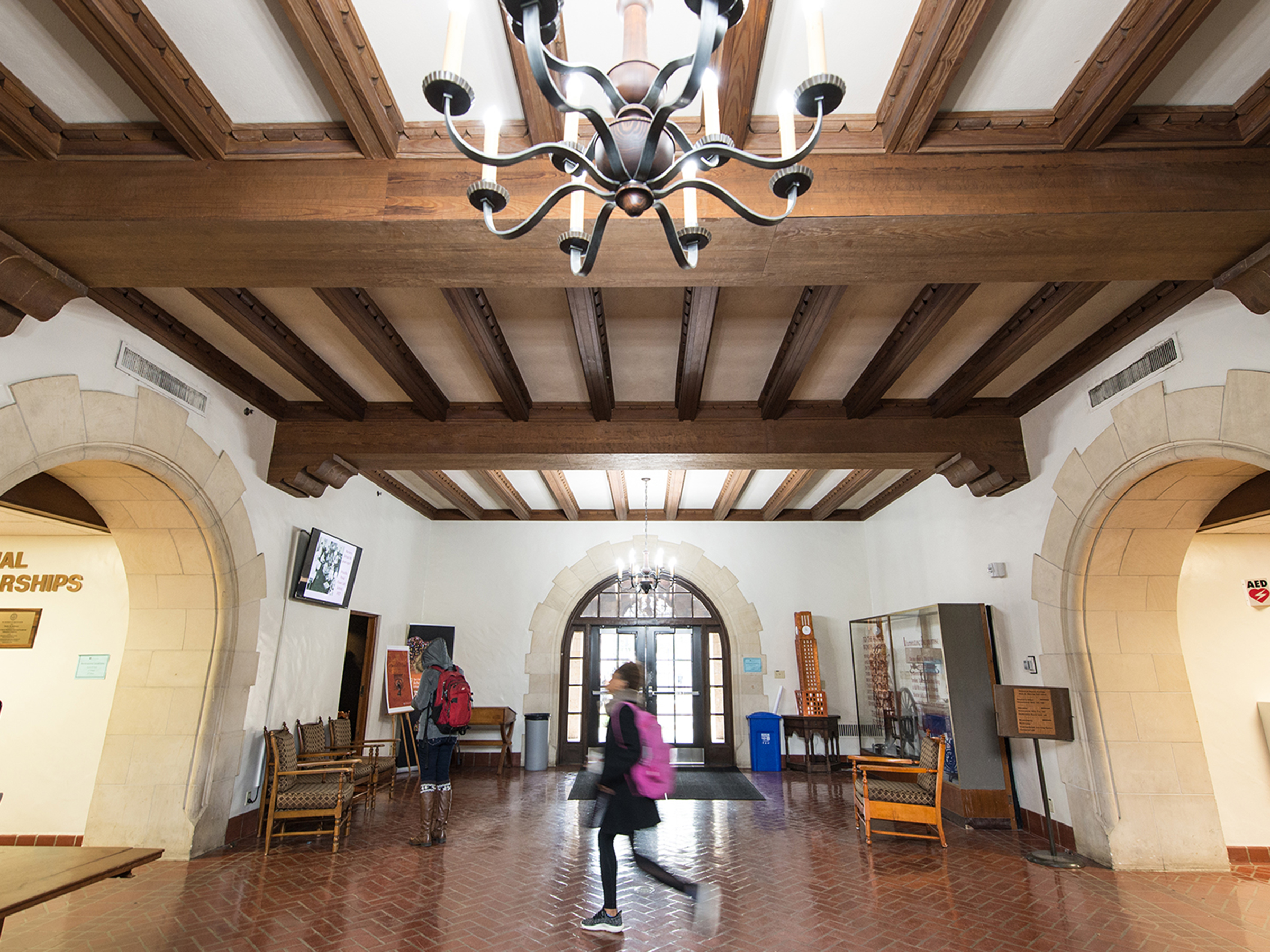 Photo of main hallway in the historic Gearing Hall. An antique chandelier frames the top amongst vaulted ceilings; below a student walks between two arched, cased-opening doorways.  