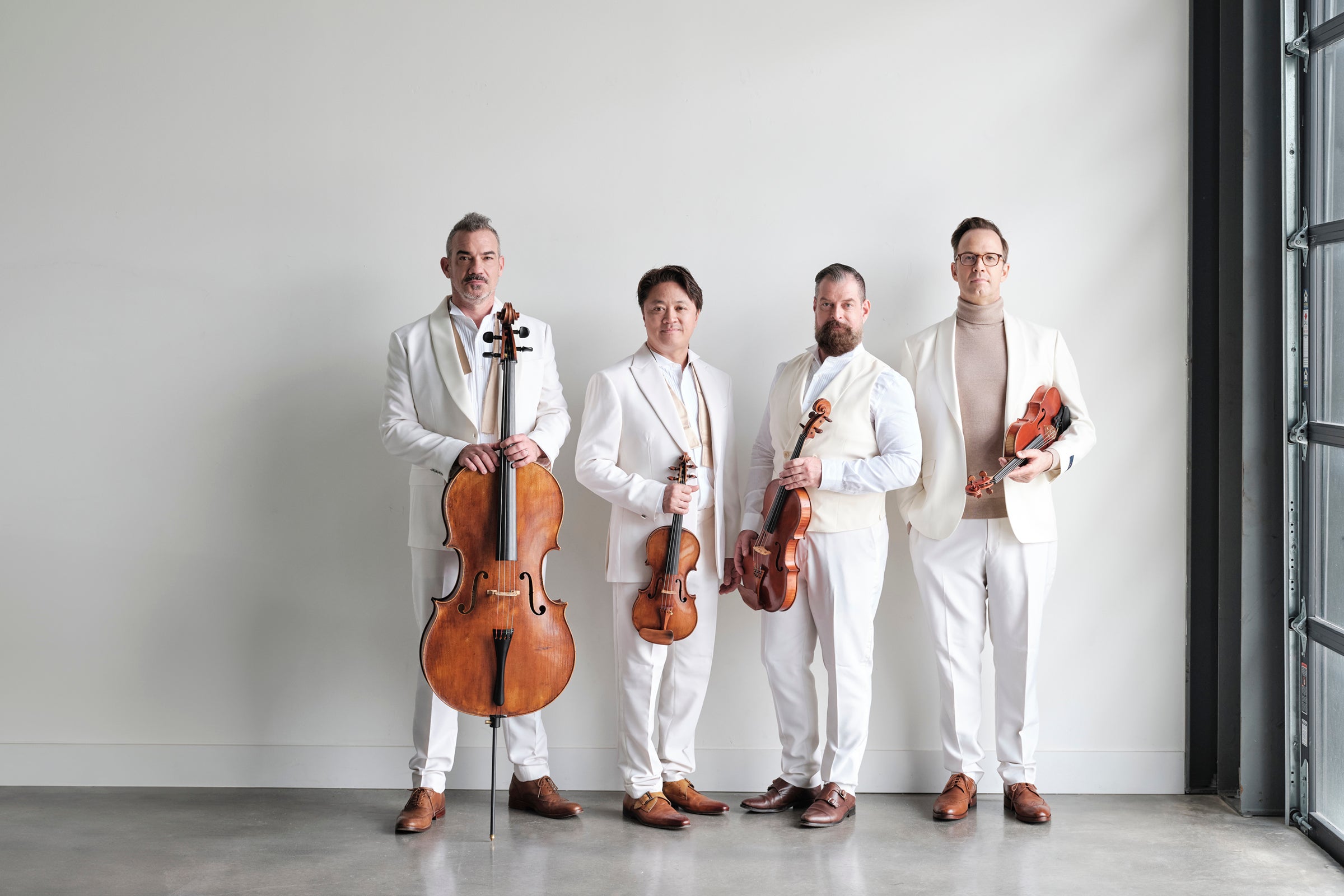 Four men in white suits stand against a wall holding various stringed instruments