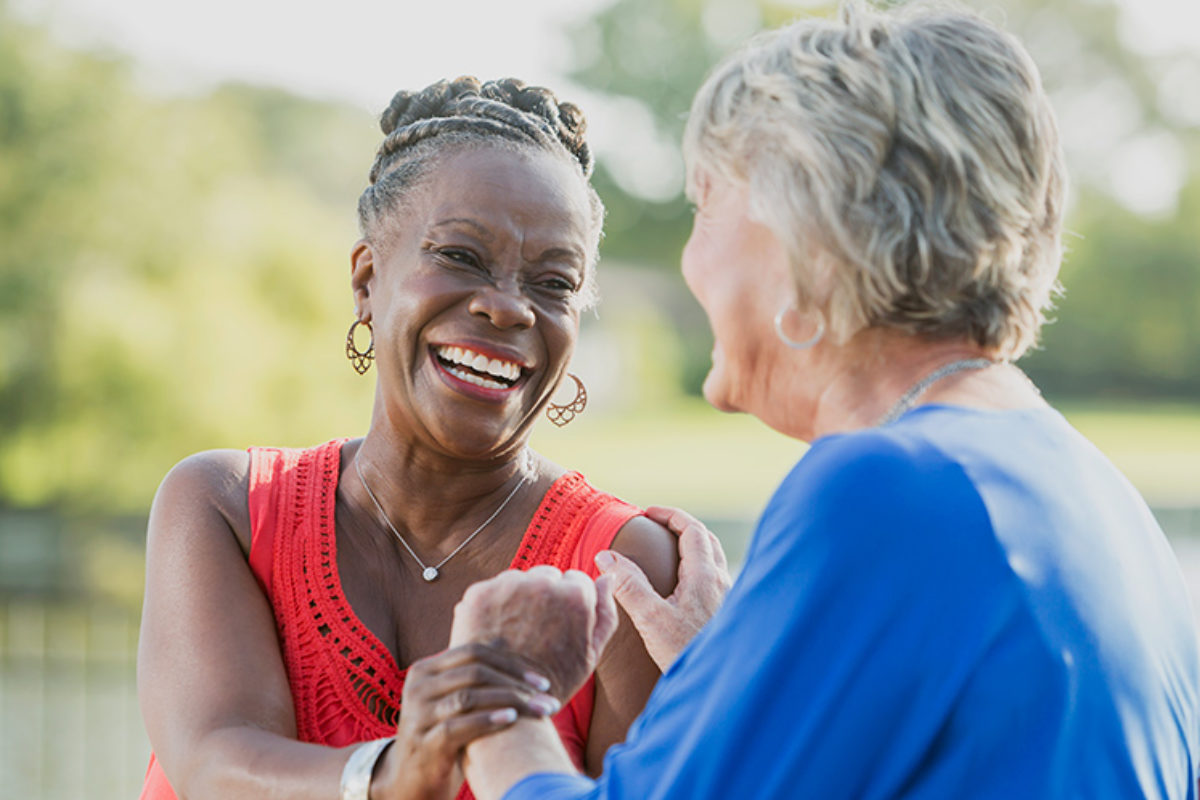 Image shows an older Black woman on the left holding hands and talking with an older White woman on the right while smiling