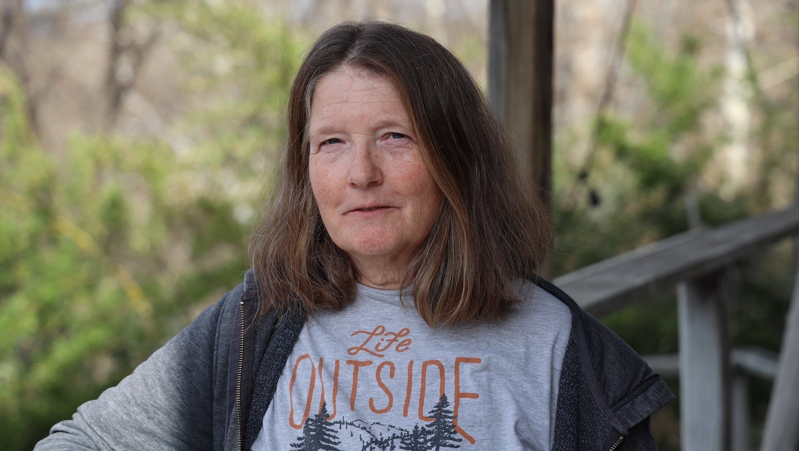 A woman dressed informally stands on a porch with greenery in the background