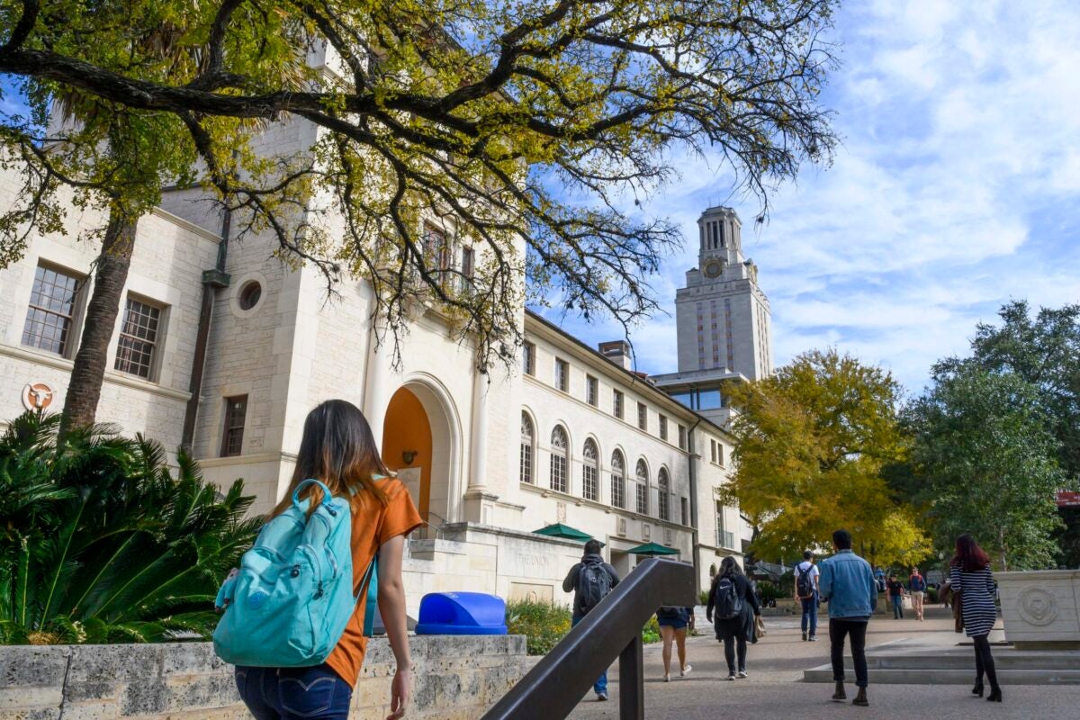 Students in backpacks walk by a limestone building as the UT Tower looms in the distance.