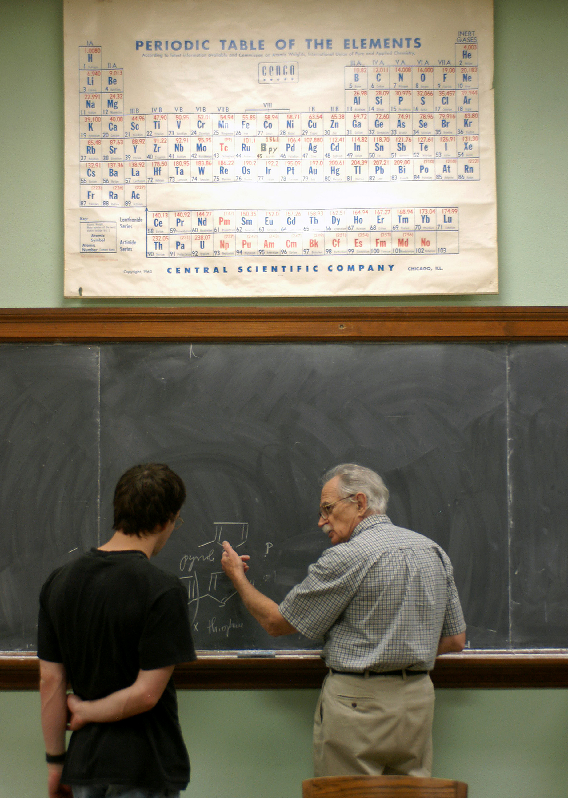 A professor and student stand at a chalkboard working on a chemistry problem