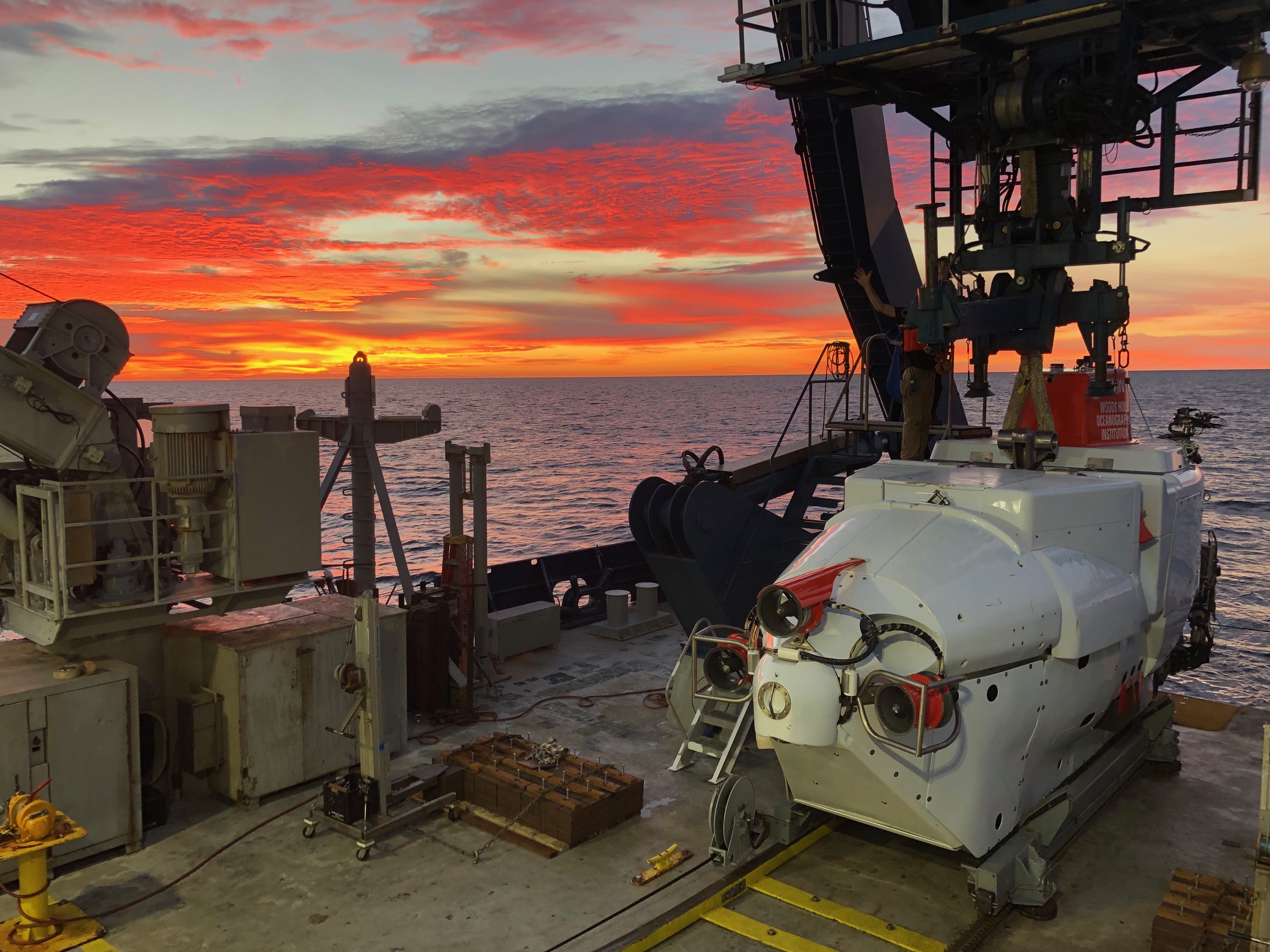 A white submersible sits on the deck of a research ship as red and orange clouds over the sea and on the horizon foretell the rising sun