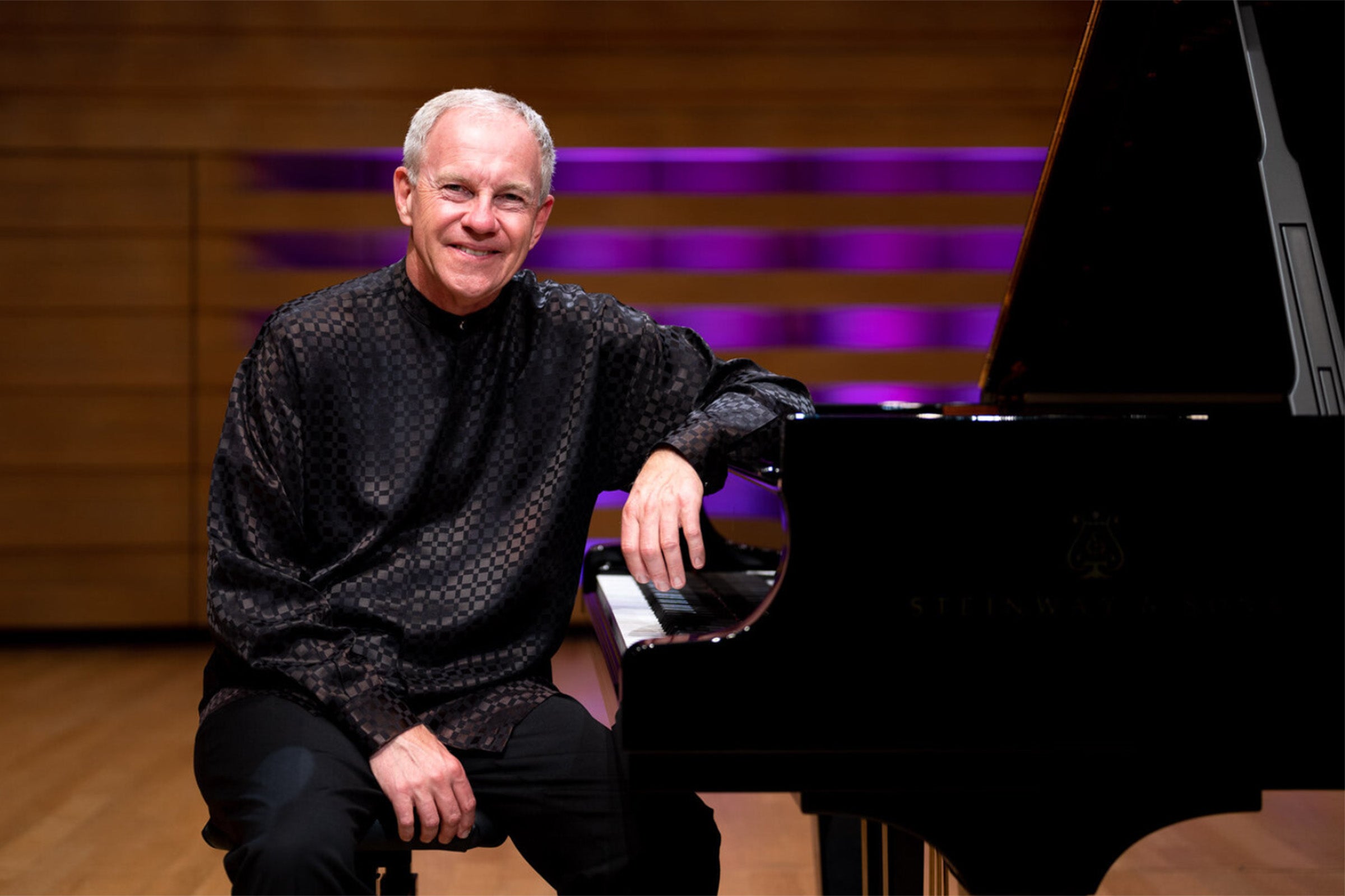 Anton Nel in a dark shirt and pants smiling while leaning on a grand piano.