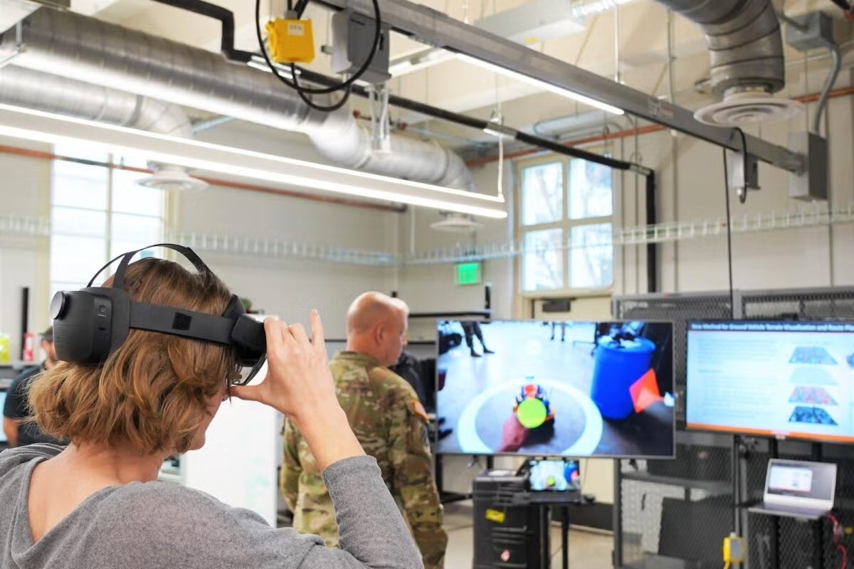 A woman in a VR headset looks at a screen while a person in miltiary camouflage looks at a larger screen in a robotics facility.