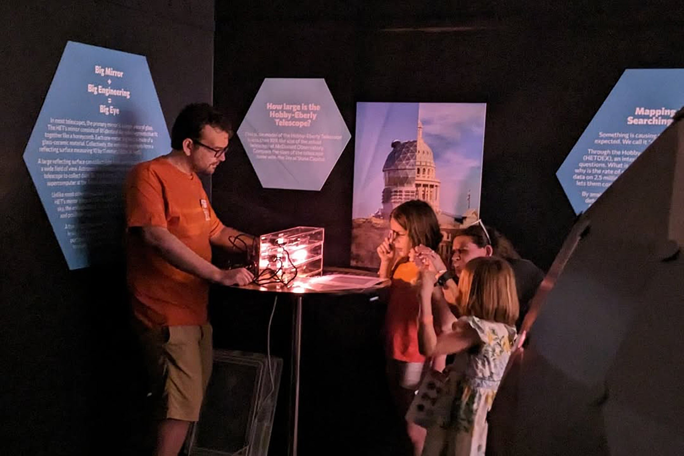 A man in a burnt orange t-shirt demonstrates spectroscopy to a father and his two daughters.