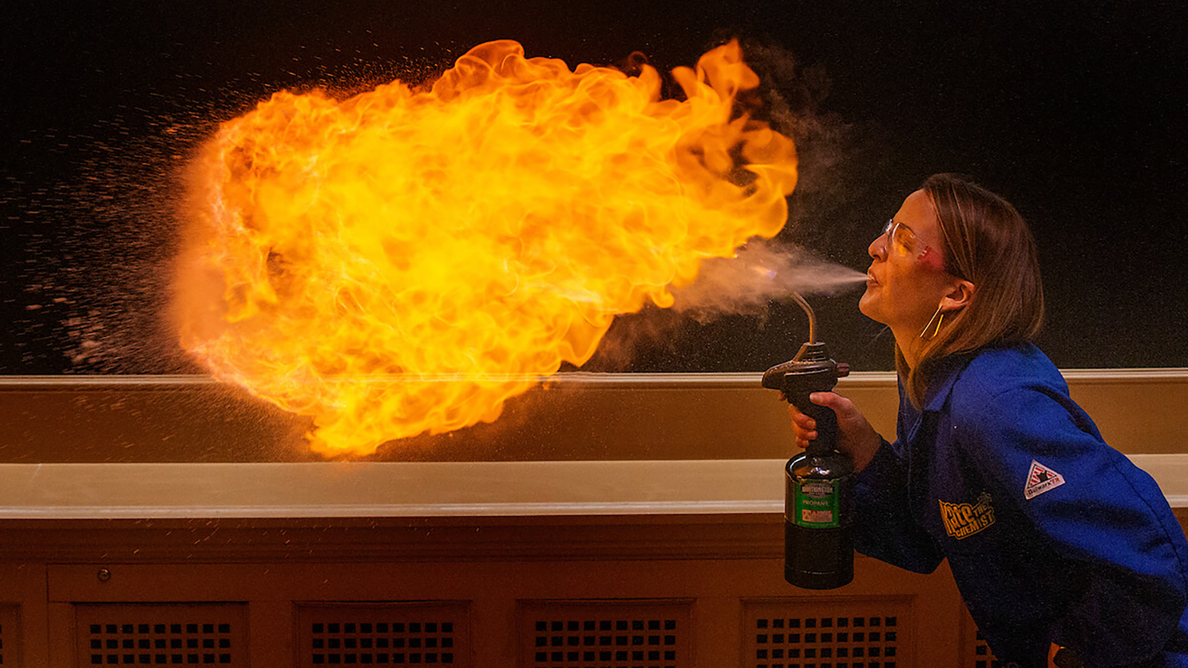 A woman in blue lab coat blows a puff of fire out of her mouth