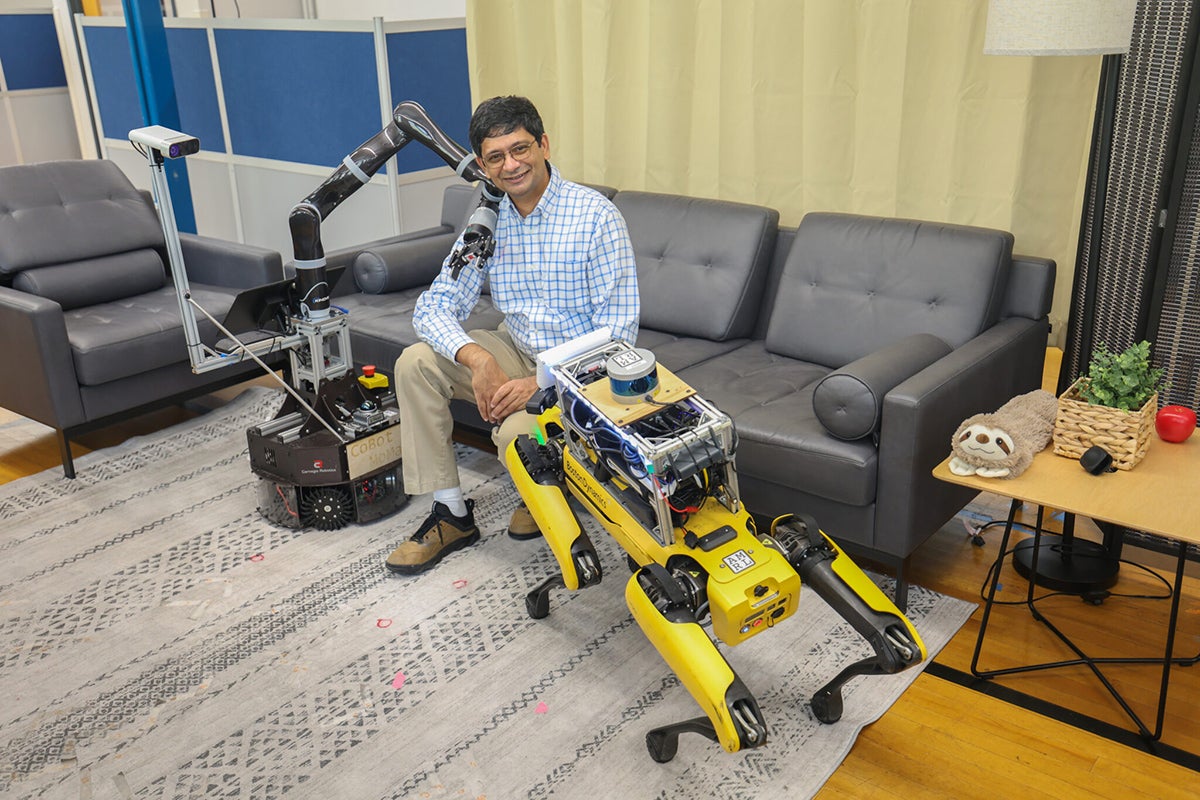 A scientist sits on a couch next to a yellow robot in the form of a dog and a gray robot with a large arm and gripper