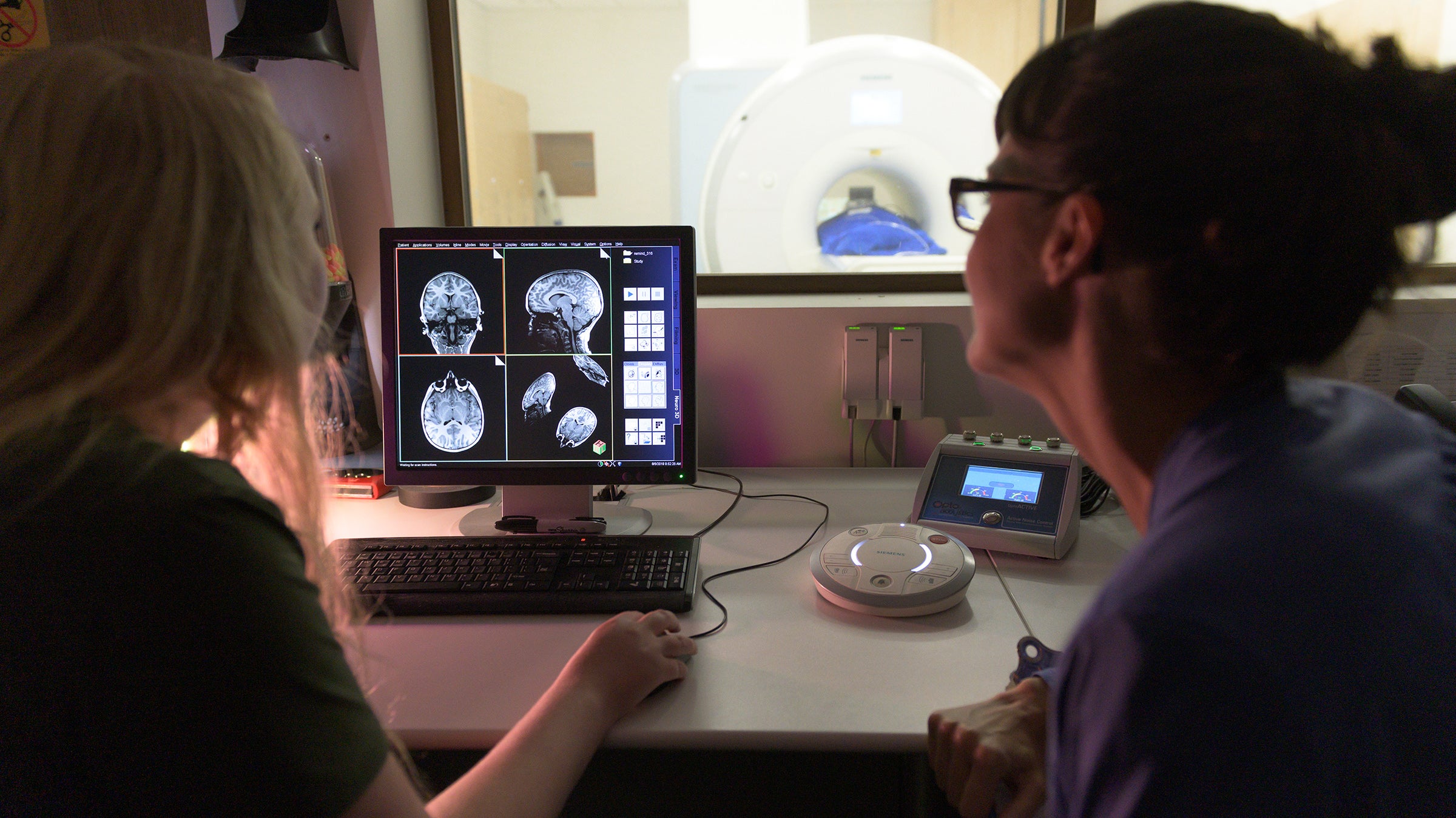 Two scientists review MRI brain scans on a screen in a control room. Through a window, we can see a patient in an MRI scanner.