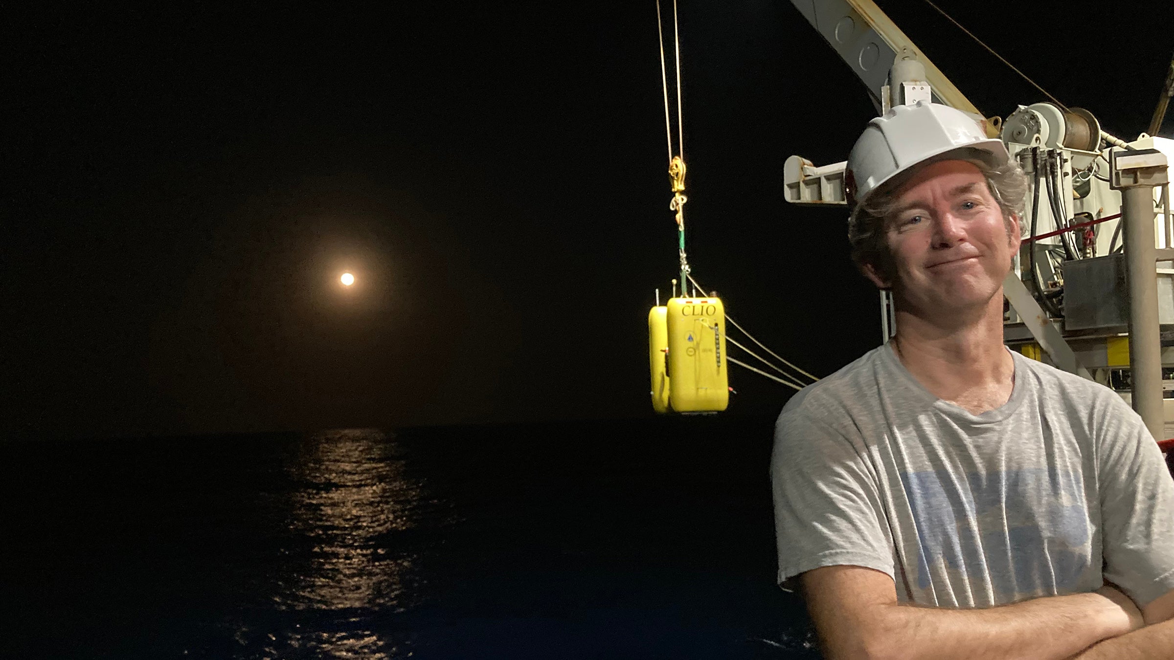 A scientist wearing a hardhad stands on the deck of a ship while a crane in the background lowers a yellow robot into the water