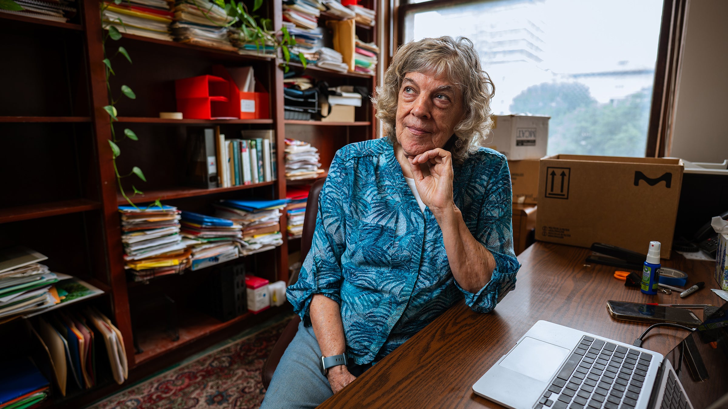 A scientist and teacher sits at her desk, behind her wooden shelves overflow with papers and books