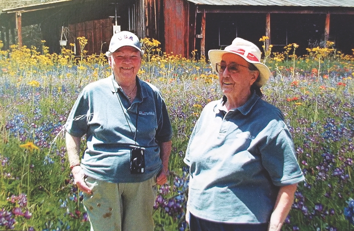 Lorraine Wyer and Casey Stengl stand in hats in an outdoor area at Stengl Lost Pines BIological Station with wildflowers in the foreground and a building farther away