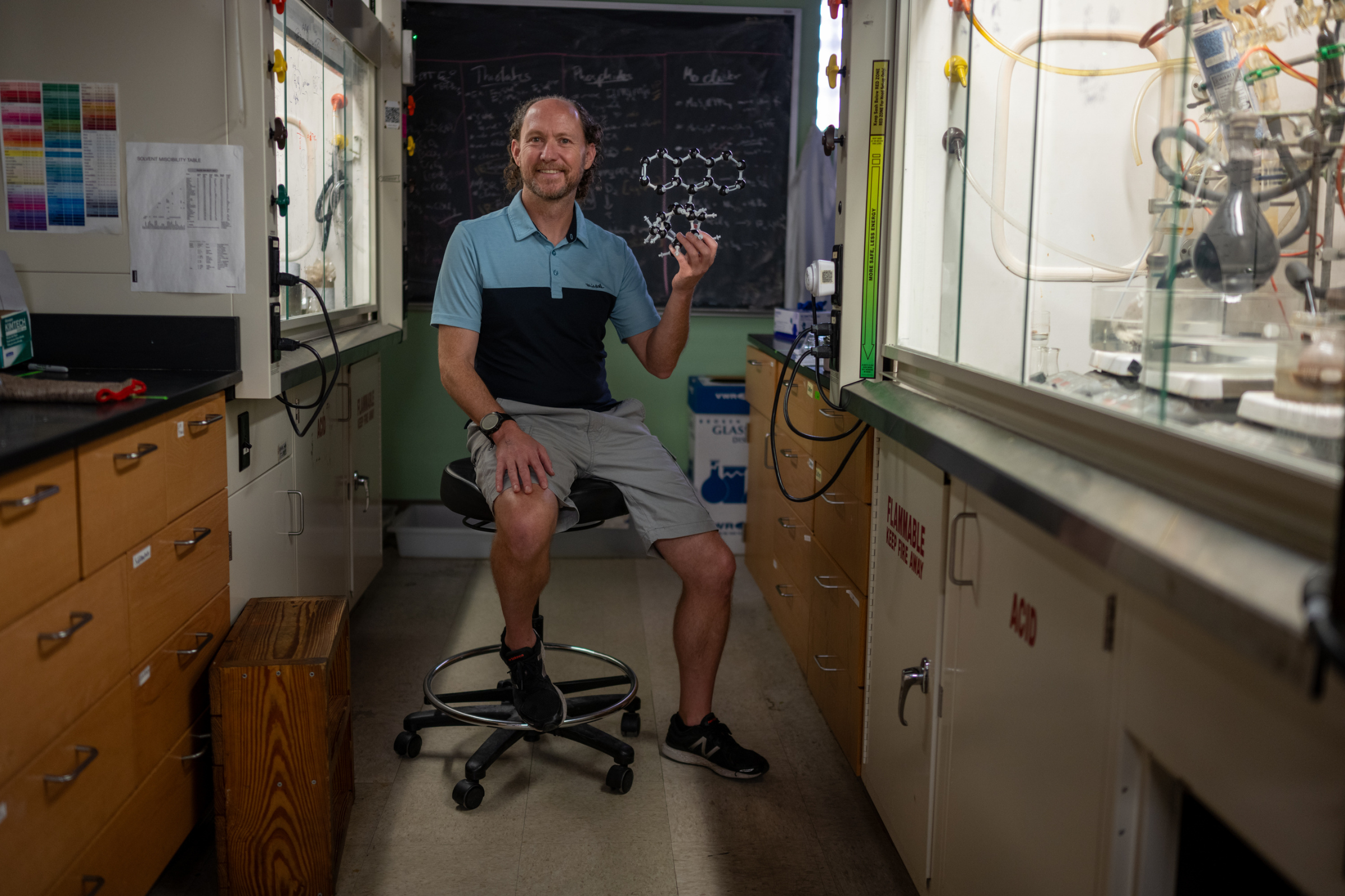 Michael Rose sits between two lab benches on a rolling stool holding a model of a molecule