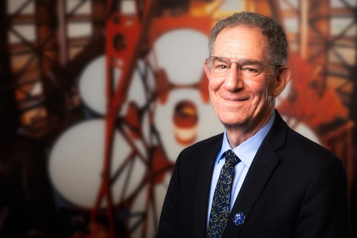 A man in a suit with a pin that looks like a multi-mirror telescope stands in front of a large model of the same telescope.
