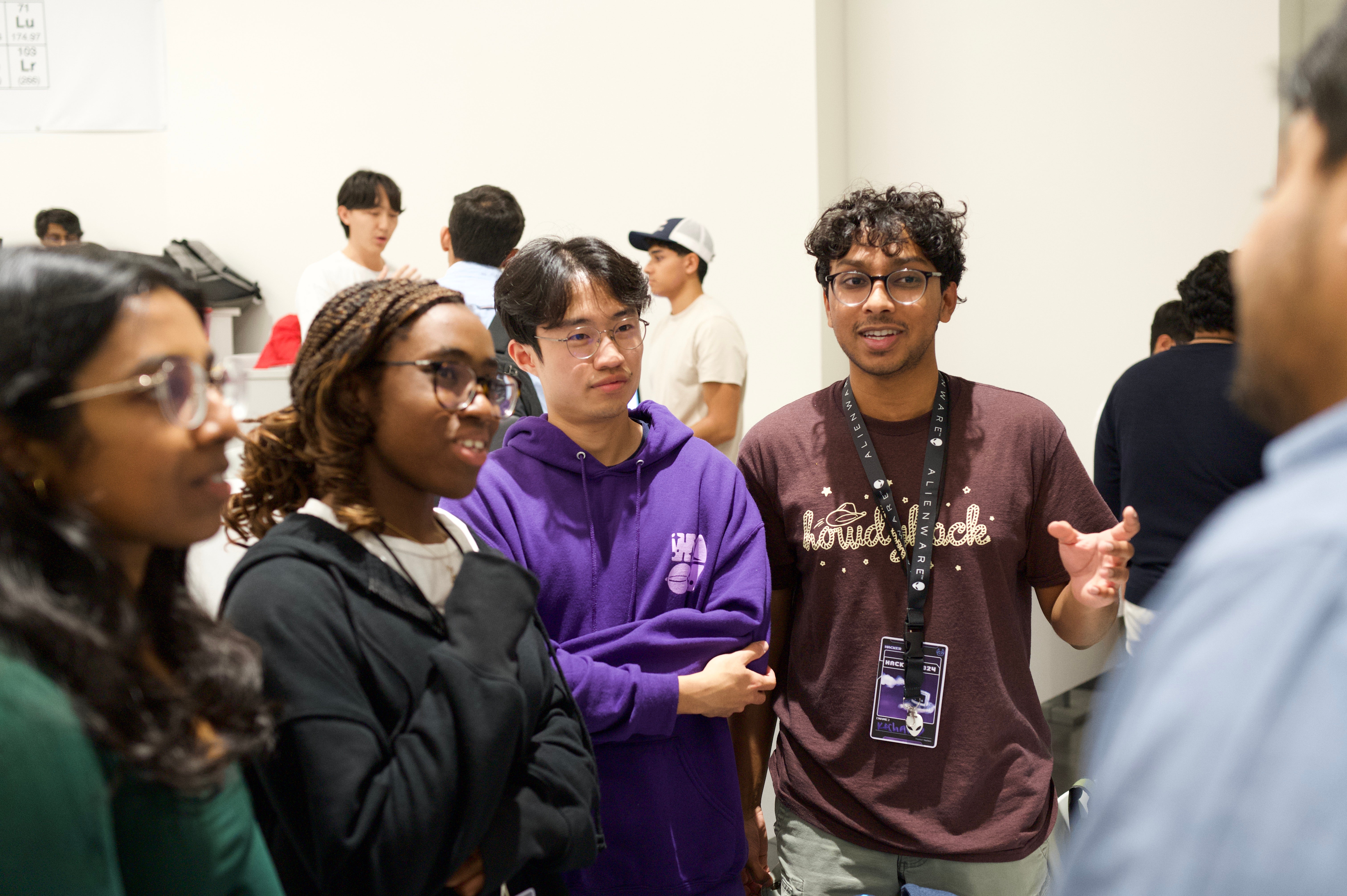 A group of five students at a hackathon discuss a problem, while other groups confer in the background.