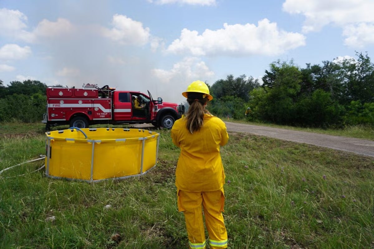 A firefighter stands near a trough of water by a pastoral road while other firefighters in and out of a small truck work.
