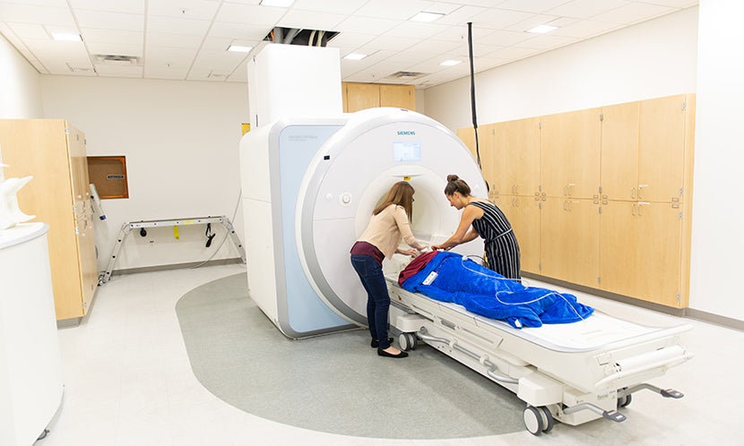 Two scientists prepare a patient to go into an fMRI scanner. The patient is laying on a horizontal structure that slides into a round opening in a large, white piece fMRI scanner.