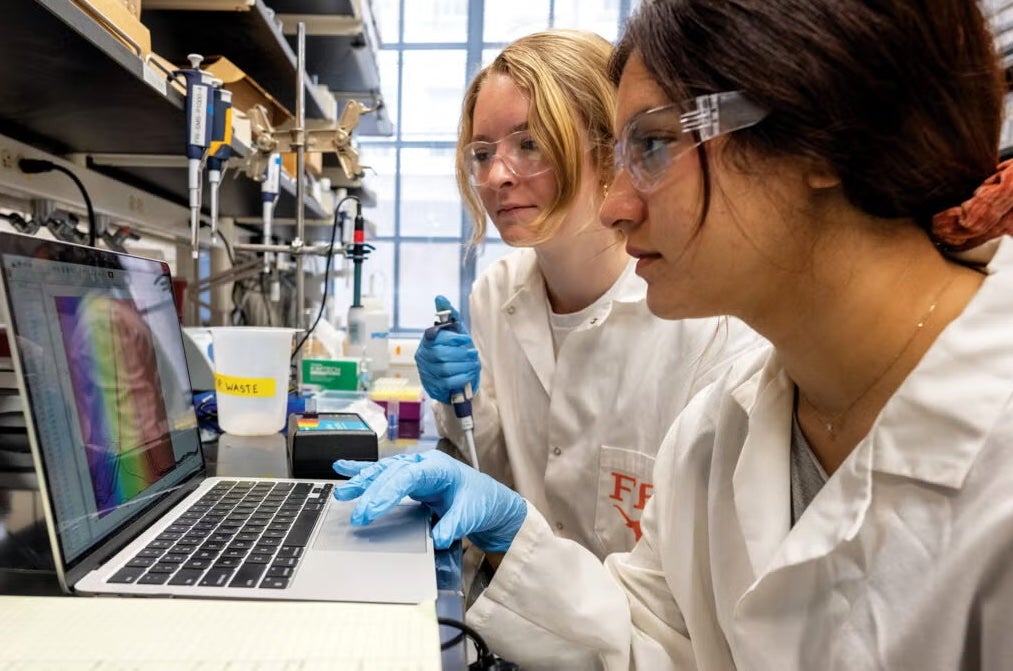 A pair of students in lab coats, goggles and gloves hold a pipette and look at a readout on a computer screen.