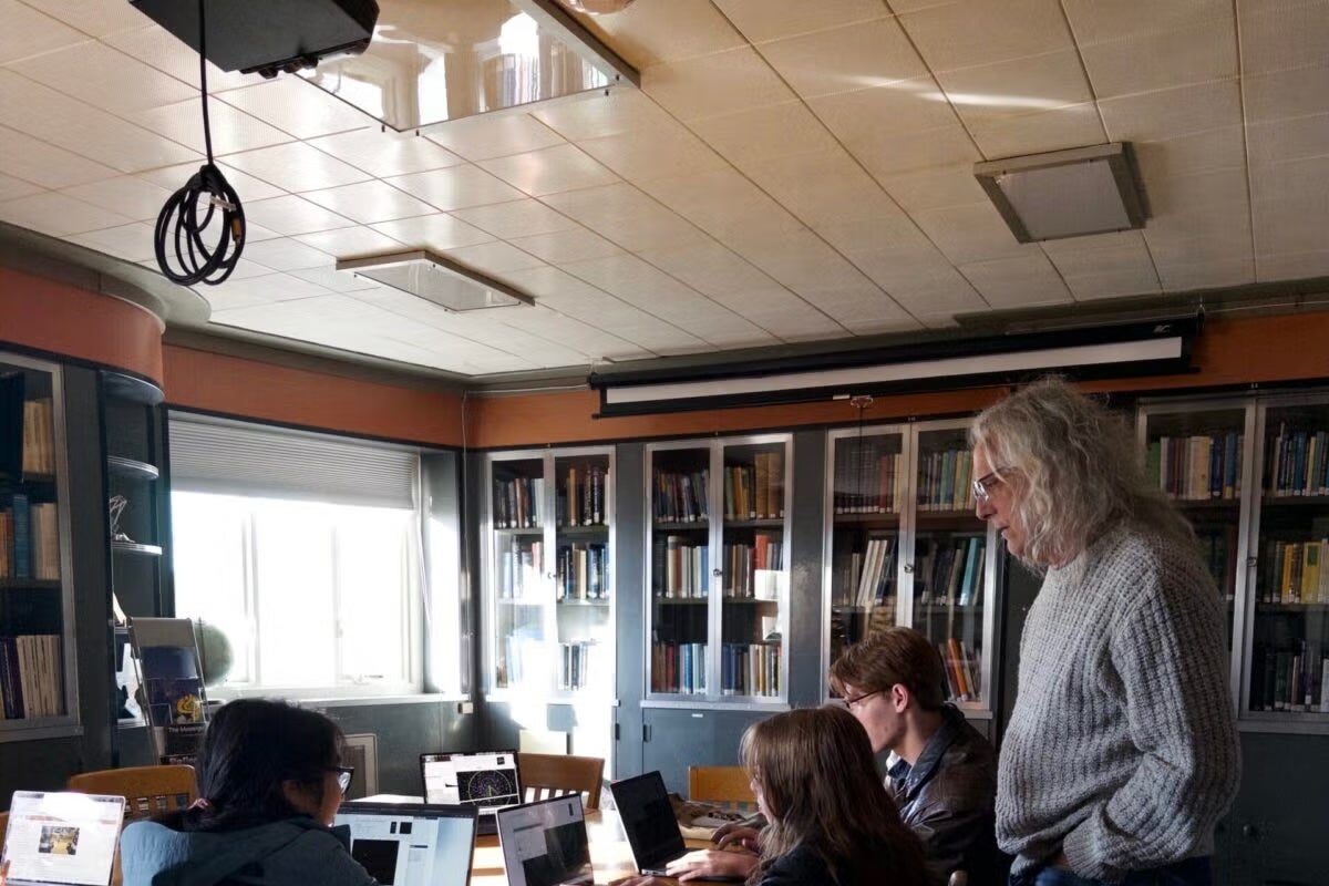 In a room lined with bookshelves, students work on their laptops as a professor, hands in pockets, guides their work.