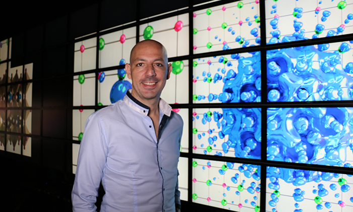 Giustino stands in front of a wall of LCD screens displaying computational models