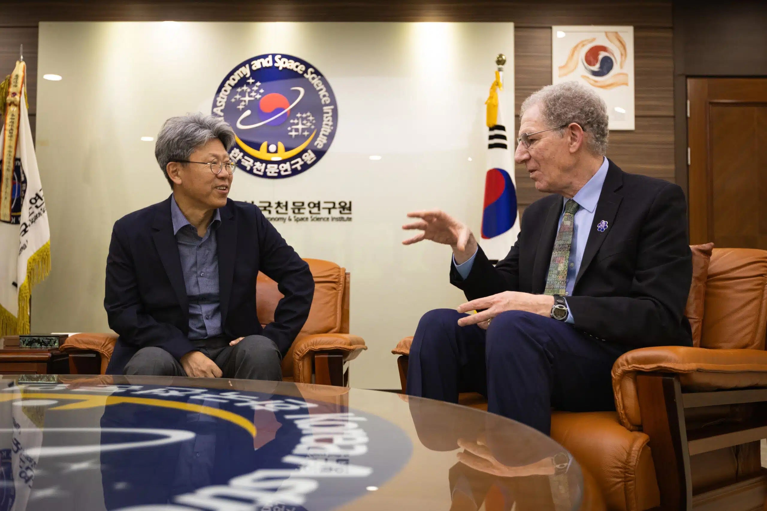 Two men in suits sit in leather chairs in a room decorated with the flag of South Korea and signage that indicates an Astronomy and Space Science Institute abroad