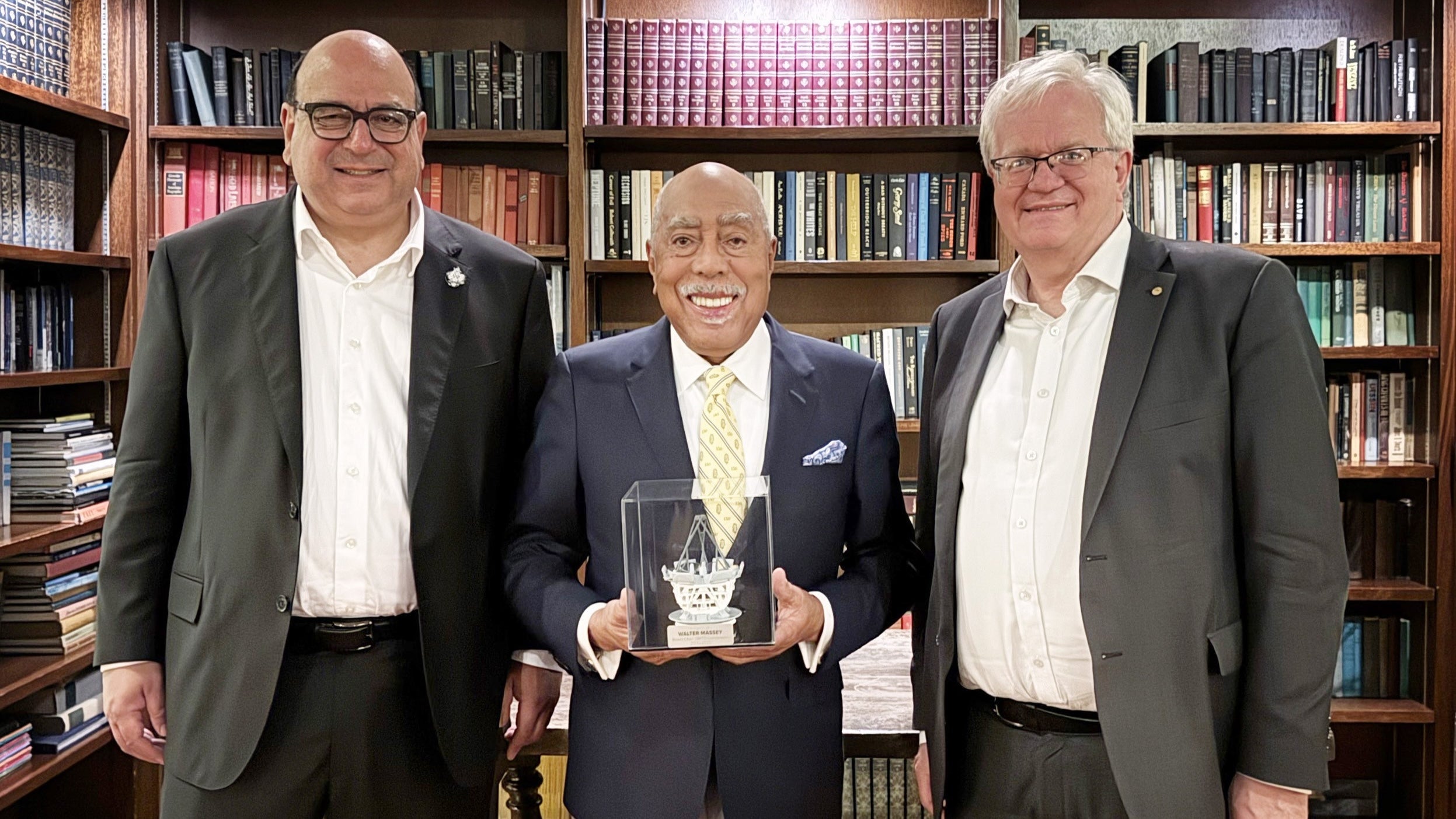 Three men in suits smile in a room lined with book-filled bookshelves, while one of the men holds a cube with a representation of a unique telescope.