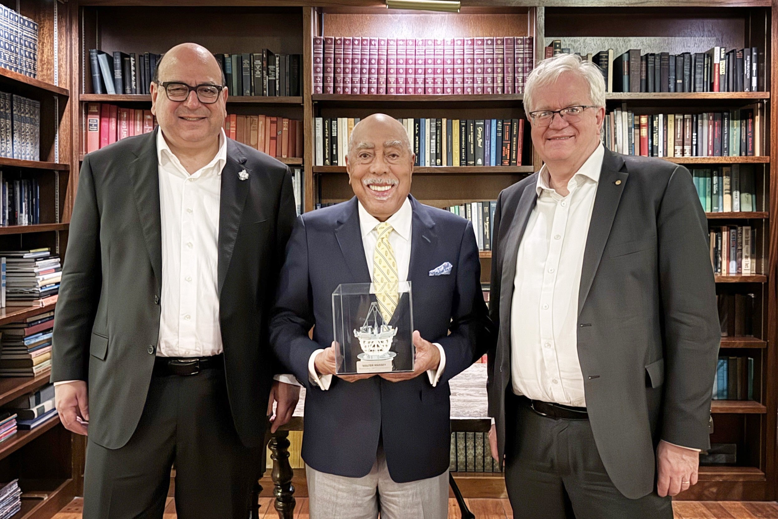 Three men in suits smile in a room lined with book-filled bookshelves, while one of the men holds a cube with a representation of a unique telescope.