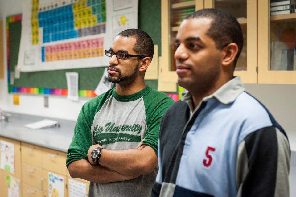 Identical twins, one with a beard and glasses, stand together in a classroom with a periodic table on the wall